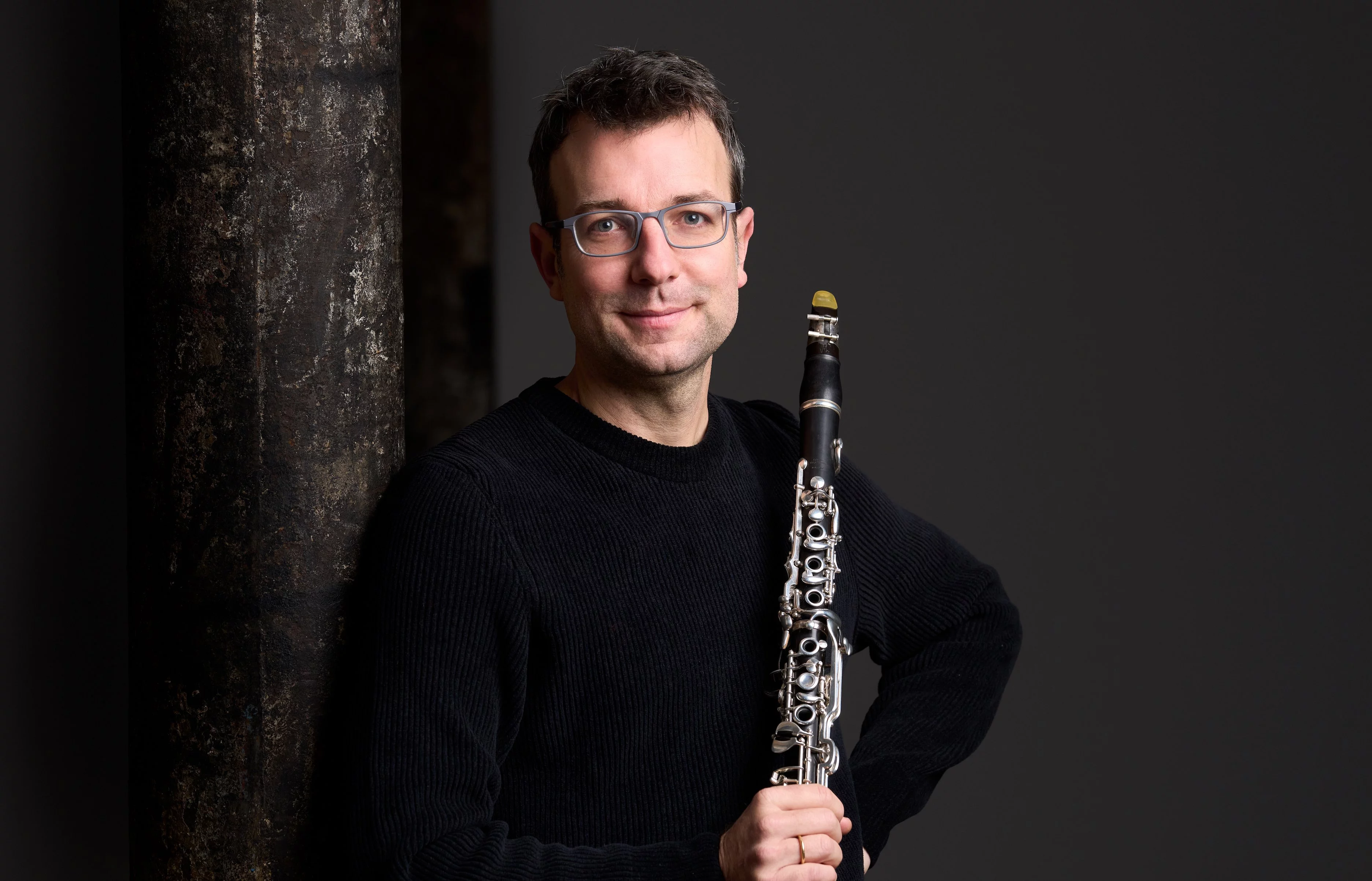 A man wearing glasses and a black sweater stands indoors, leaning against a rusty metal pillar and holding a clarinet, with a neutral gray background.