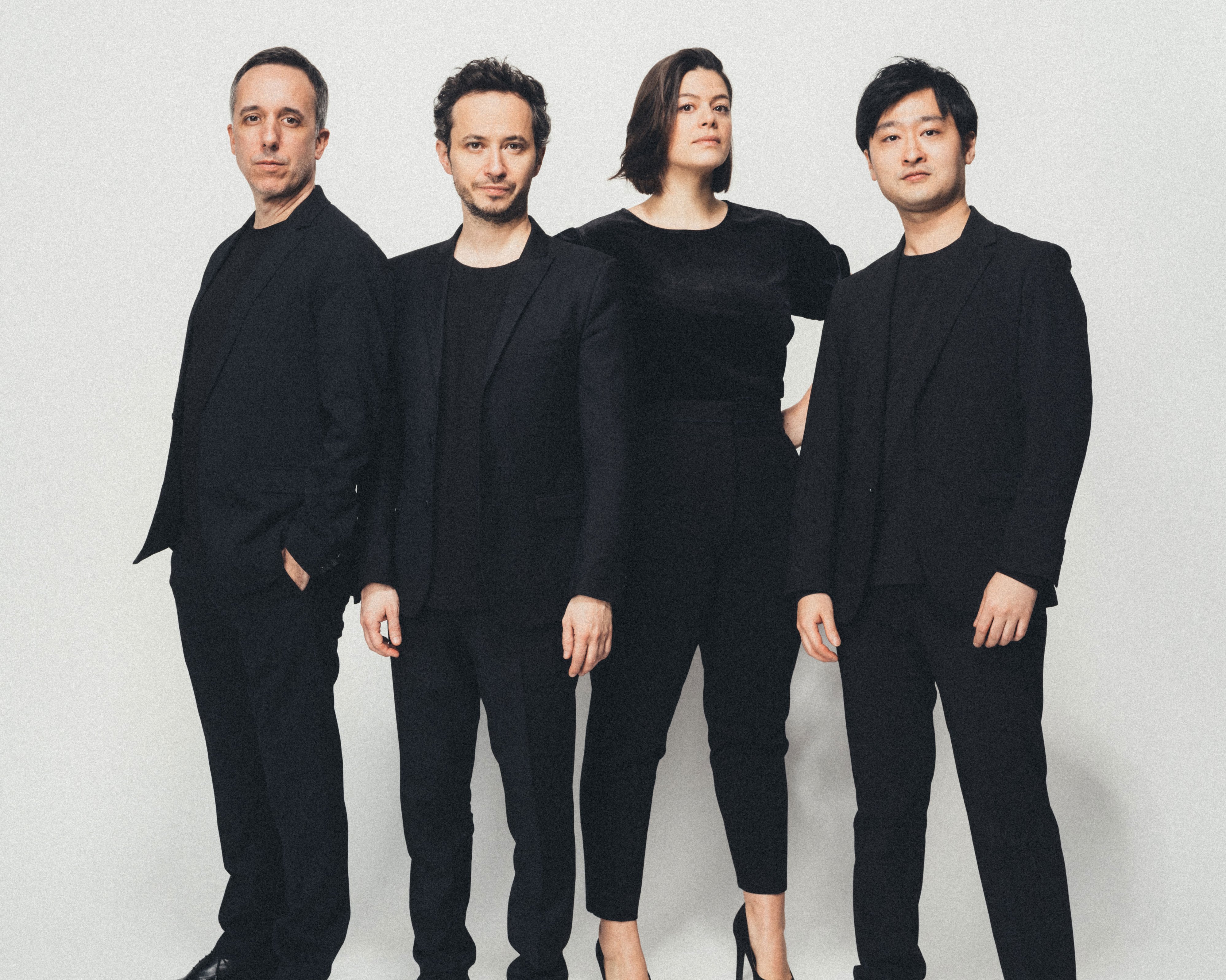 Vier Personen stehen vor einem schlichten weißen Hintergrund, alle in schwarzer Kleidung gekleidet. Sie blicken mit neutralem Gesichtsausdruck nach vorne, und ihre Posen wirken selbstbewusst und professionell.