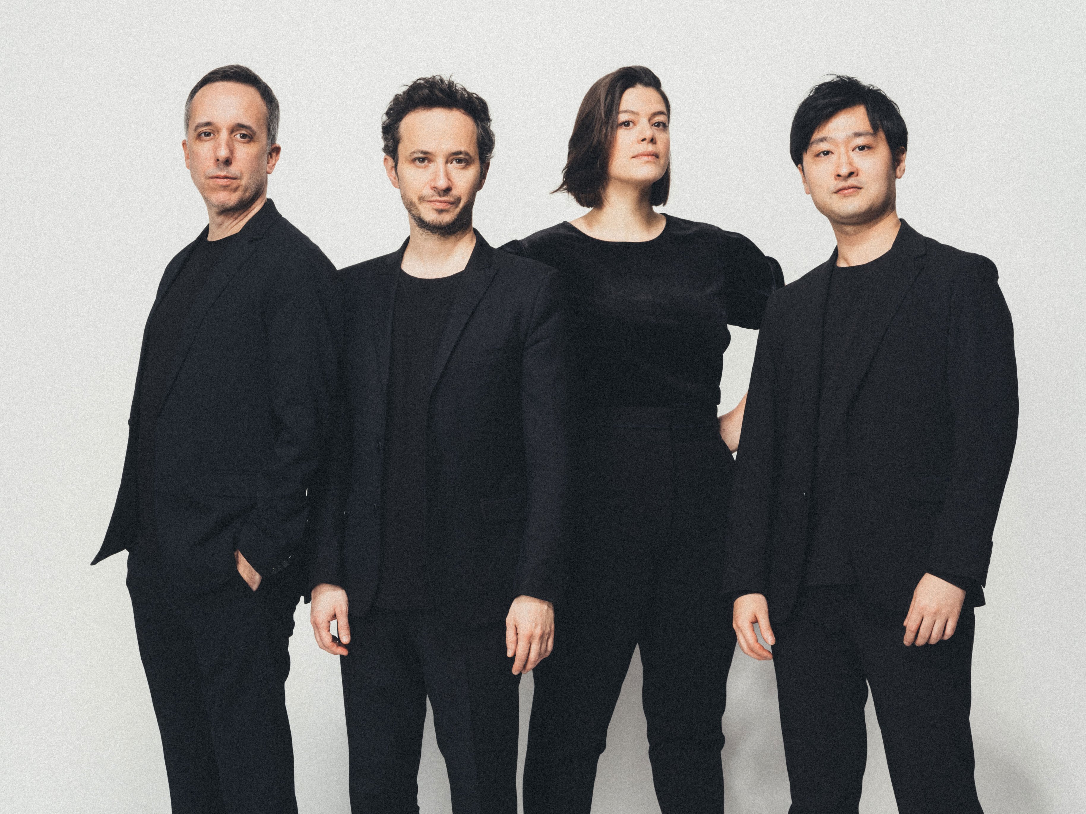 Vier Personen stehen vor einem schlichten weißen Hintergrund, alle in schwarzer Kleidung gekleidet. Sie blicken mit neutralem Gesichtsausdruck nach vorne, und ihre Posen wirken selbstbewusst und professionell.