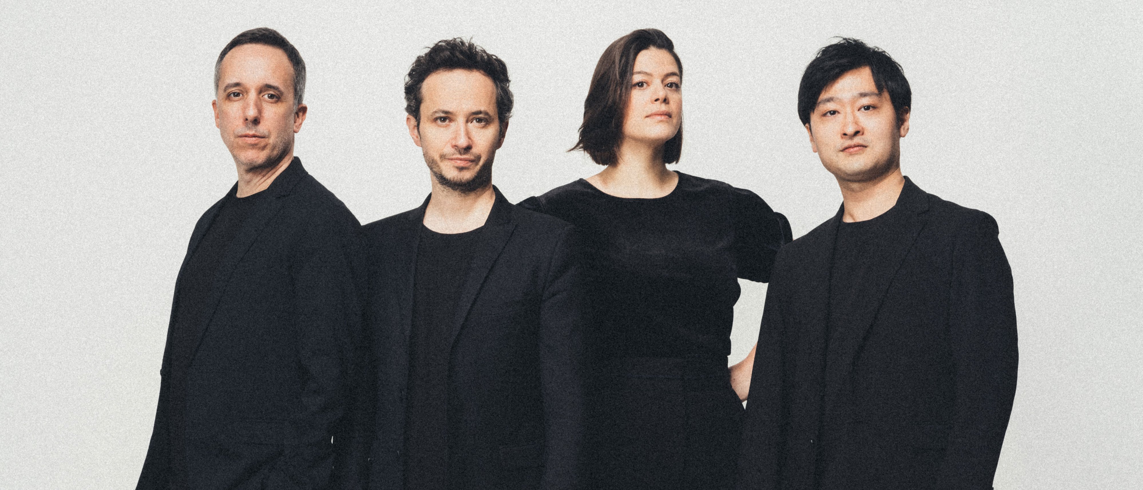 Vier Personen stehen vor einem schlichten weißen Hintergrund, alle in schwarzer Kleidung gekleidet. Sie blicken mit neutralem Gesichtsausdruck nach vorne, und ihre Posen wirken selbstbewusst und professionell.