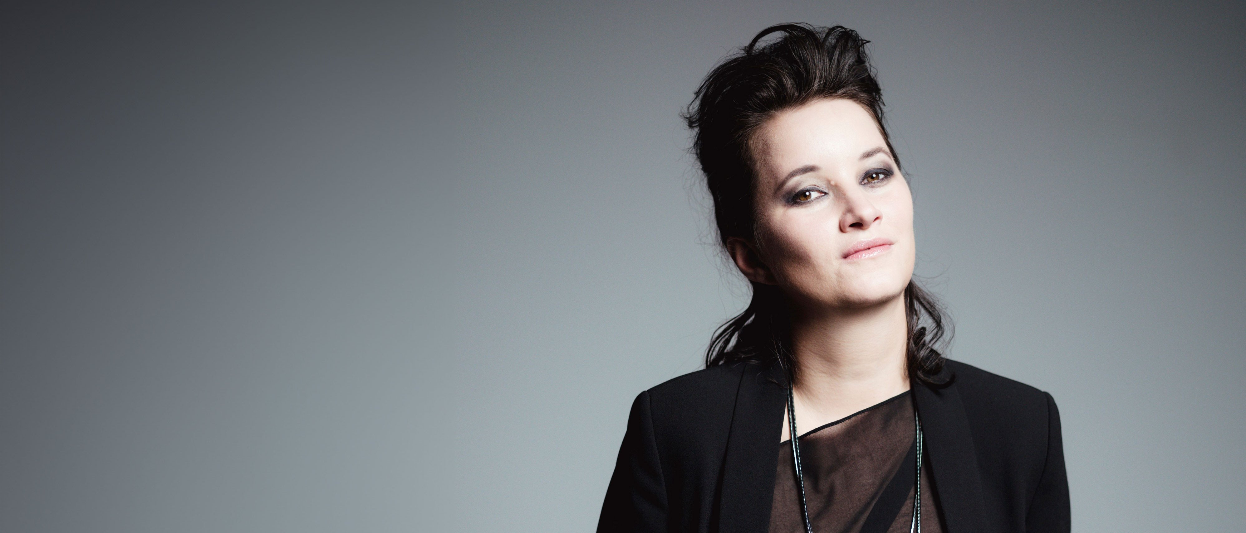 Anna Prohaska mit dunklem, gestyltem Haar und schwarzem Outfit posiert selbstbewusst vor einem schlichten grauen Hintergrund.