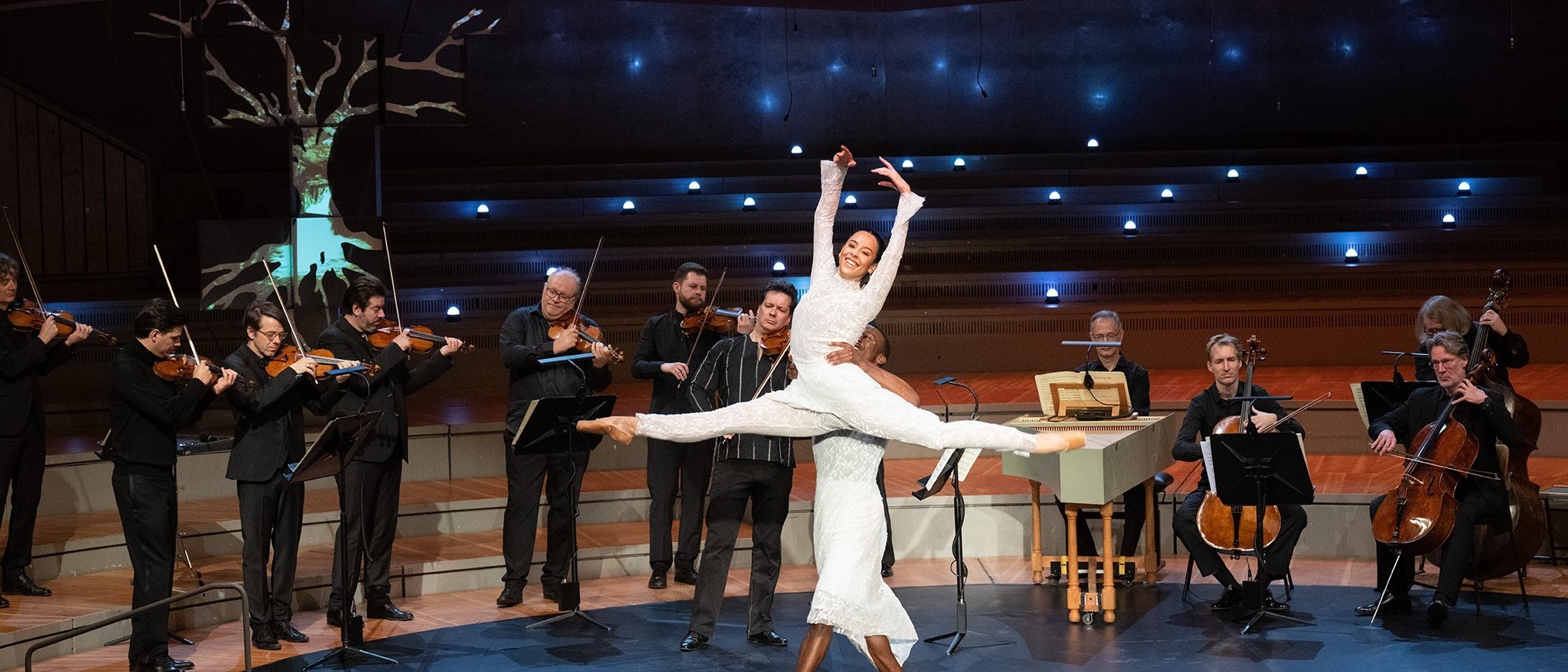 Eine Ballerina in einem weißen Outfit springt anmutig, während ein männlicher Tänzer sie hochhebt; im Hintergrund spielen Musiker auf einer Konzertbühne mit einer beleuchteten, baumähnlichen Requisite Streichinstrumente.