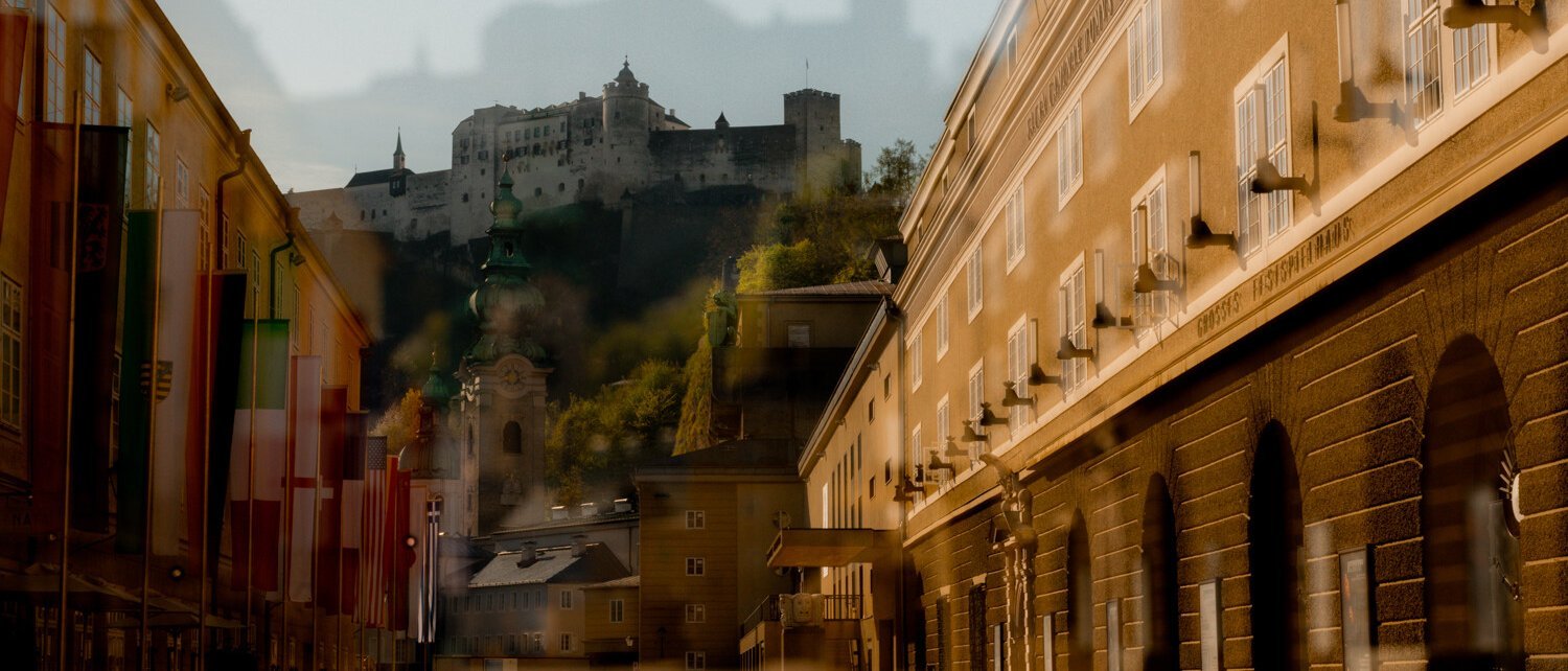 Eine sonnenbeschienene Straße in Salzburg mit Fahnen auf der linken Seite, historischen Gebäuden und der Festung Hohensalzburg auf einem Hügel im Hintergrund; das Bild hat einen Doppelbelichtungseffekt.