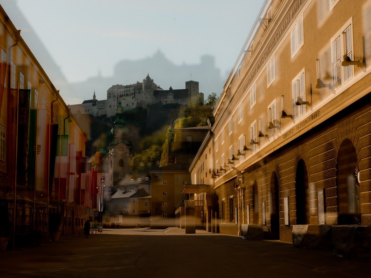 Eine sonnenbeschienene Straße in Salzburg mit Fahnen auf der linken Seite, historischen Gebäuden und der Festung Hohensalzburg auf einem Hügel im Hintergrund; das Bild hat einen Doppelbelichtungseffekt.