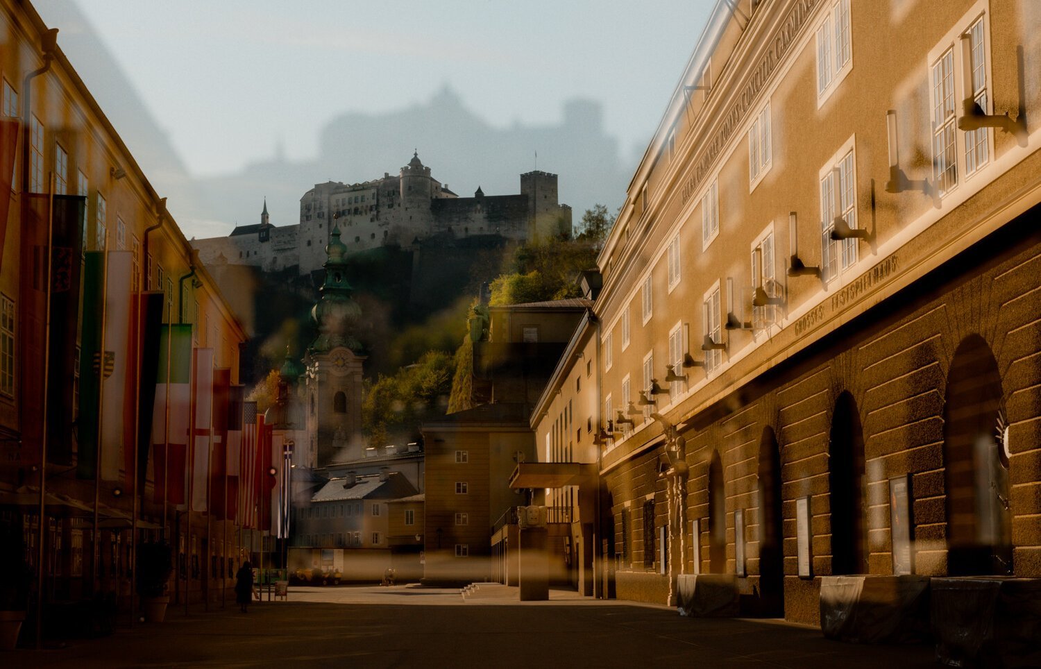 Eine sonnenbeschienene Straße in Salzburg mit Fahnen auf der linken Seite, historischen Gebäuden und der Festung Hohensalzburg auf einem Hügel im Hintergrund; das Bild hat einen Doppelbelichtungseffekt.