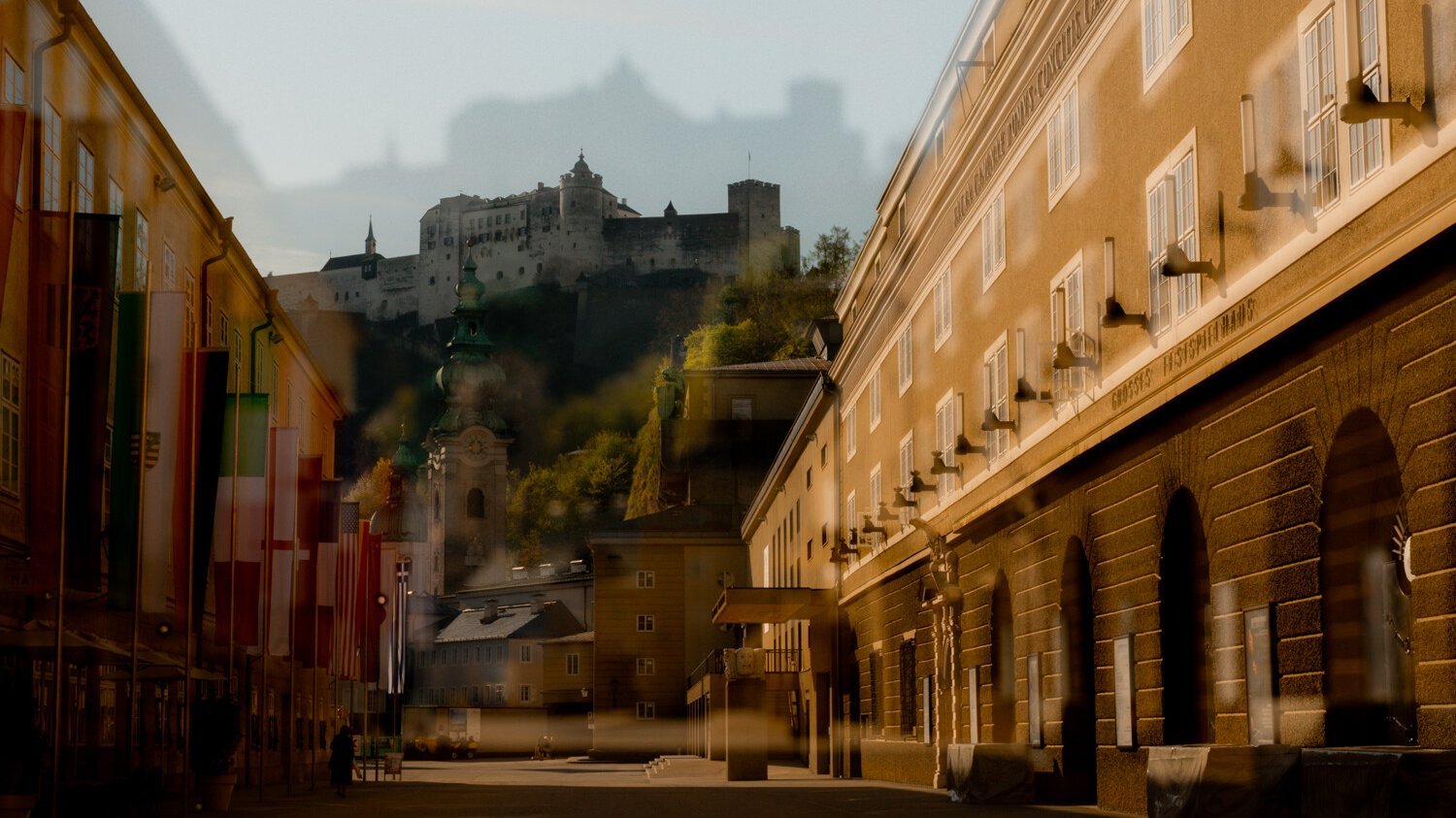 Eine sonnenbeschienene Straße in Salzburg mit Fahnen auf der linken Seite, historischen Gebäuden und der Festung Hohensalzburg auf einem Hügel im Hintergrund; das Bild hat einen Doppelbelichtungseffekt.