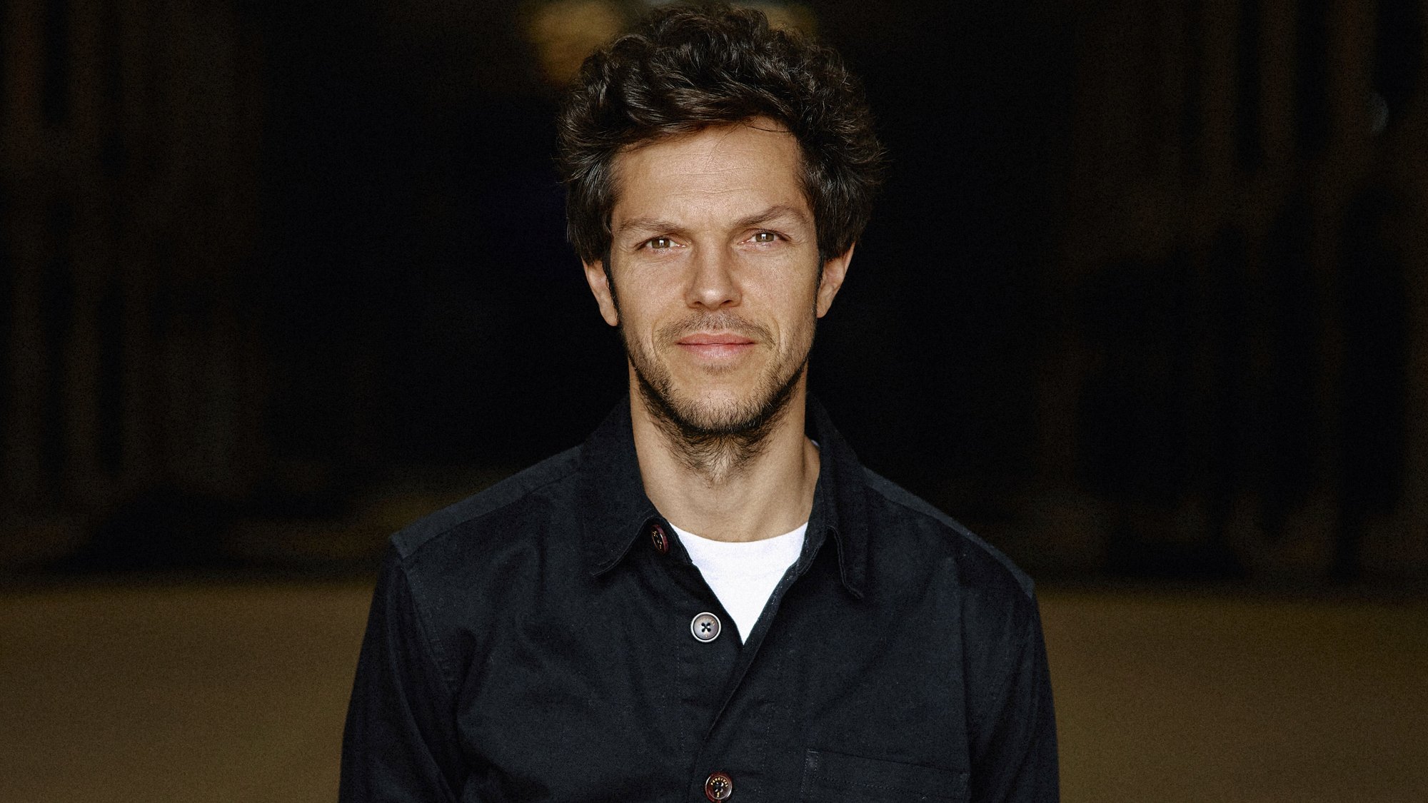 Raphael Pichon mit dunklem Haar steht in einer leeren Lagerhalle. Sie trägt eine schwarze Jacke und Jeans und lächelt leicht mit den Händen hinter dem Rücken. Der Hintergrund ist schwach beleuchtet, wodurch die Figur der Person hervorgehoben wird.