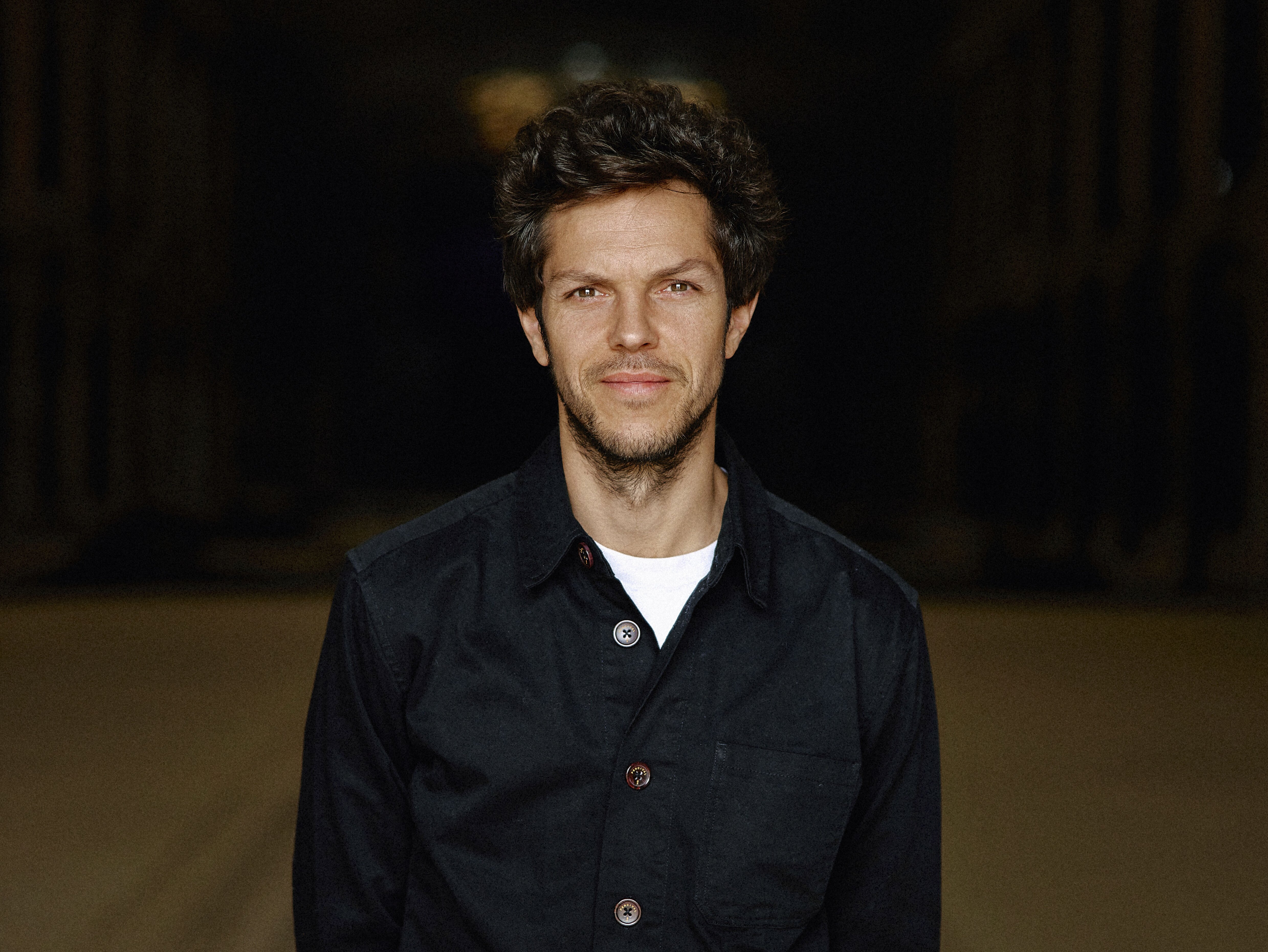 Raphael Pichon mit dunklem Haar steht in einer leeren Lagerhalle. Sie trägt eine schwarze Jacke und Jeans und lächelt leicht mit den Händen hinter dem Rücken. Der Hintergrund ist schwach beleuchtet, wodurch die Figur der Person hervorgehoben wird.