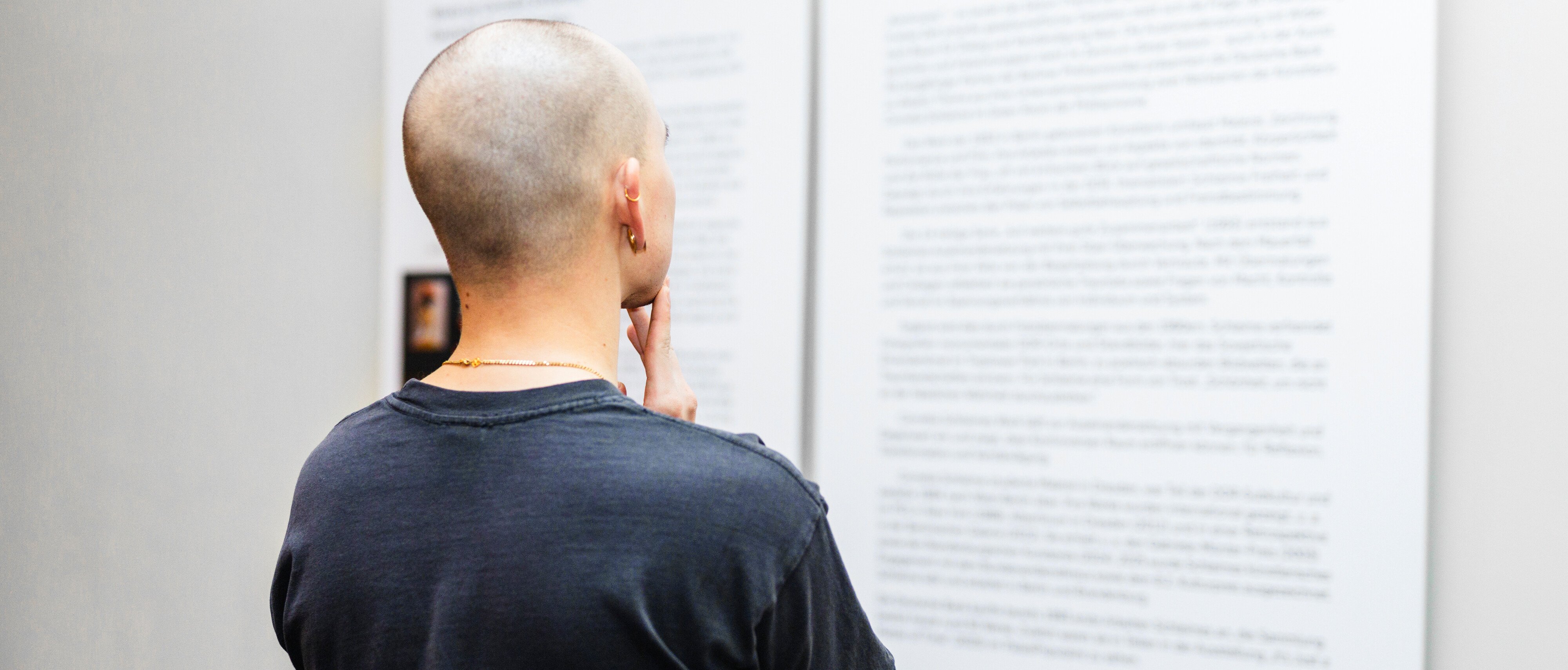 A person with a shaved head and a dark shirt stands with their back to the camera, thoughtfully reading text panels displayed on a wall in an exhibition or gallery.