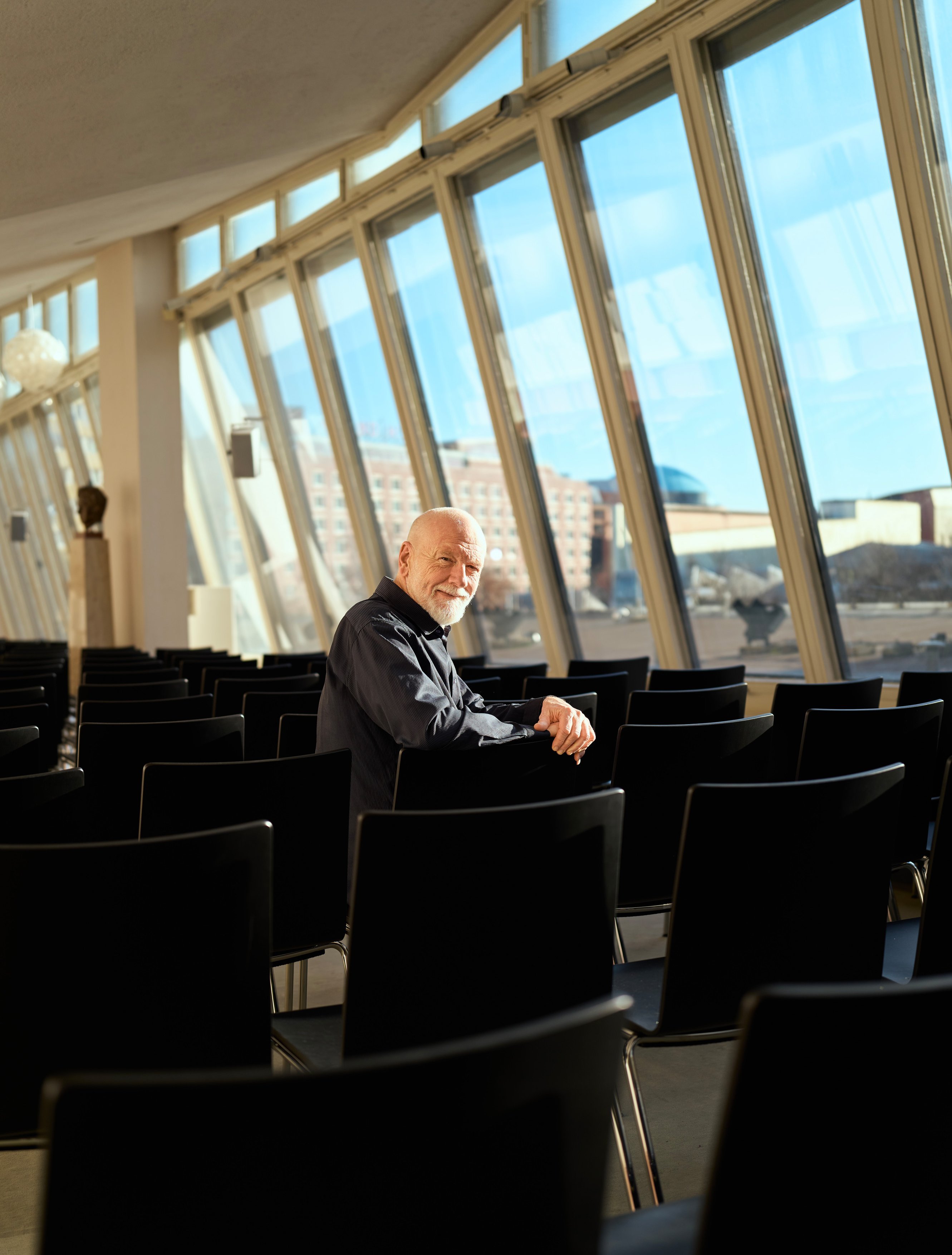 Ein älterer Mann mit weißem Bart sitzt allein zwischen Reihen schwarzer Stühle in einem sonnendurchfluteten Raum mit großen, schrägen Fenstern, die den Blick auf die Gebäude draußen freigeben. Er blickt zurück und lächelt in die Kamera.