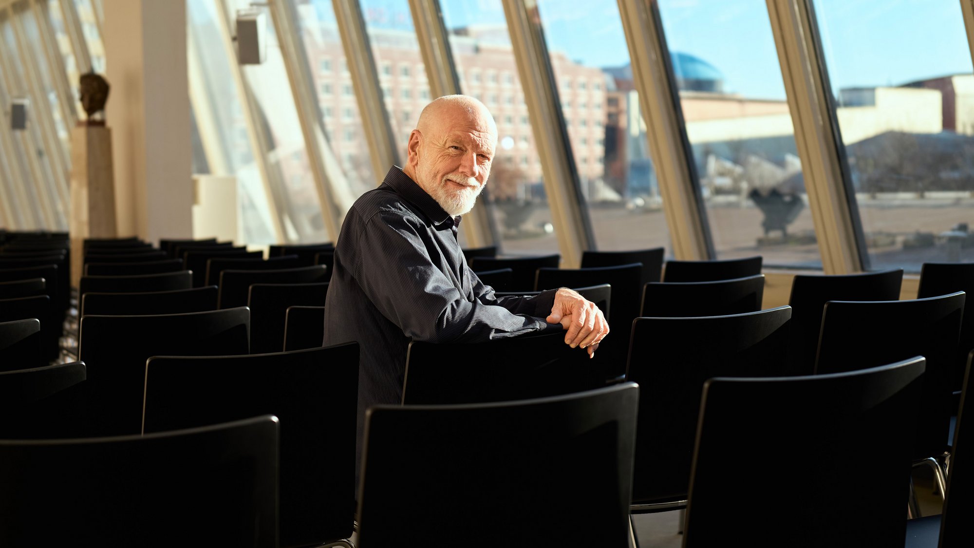 An older man with a beard sits alone in a sunlit room filled with black chairs, turning to smile at the camera. Large windows behind him reveal a cityscape outside.