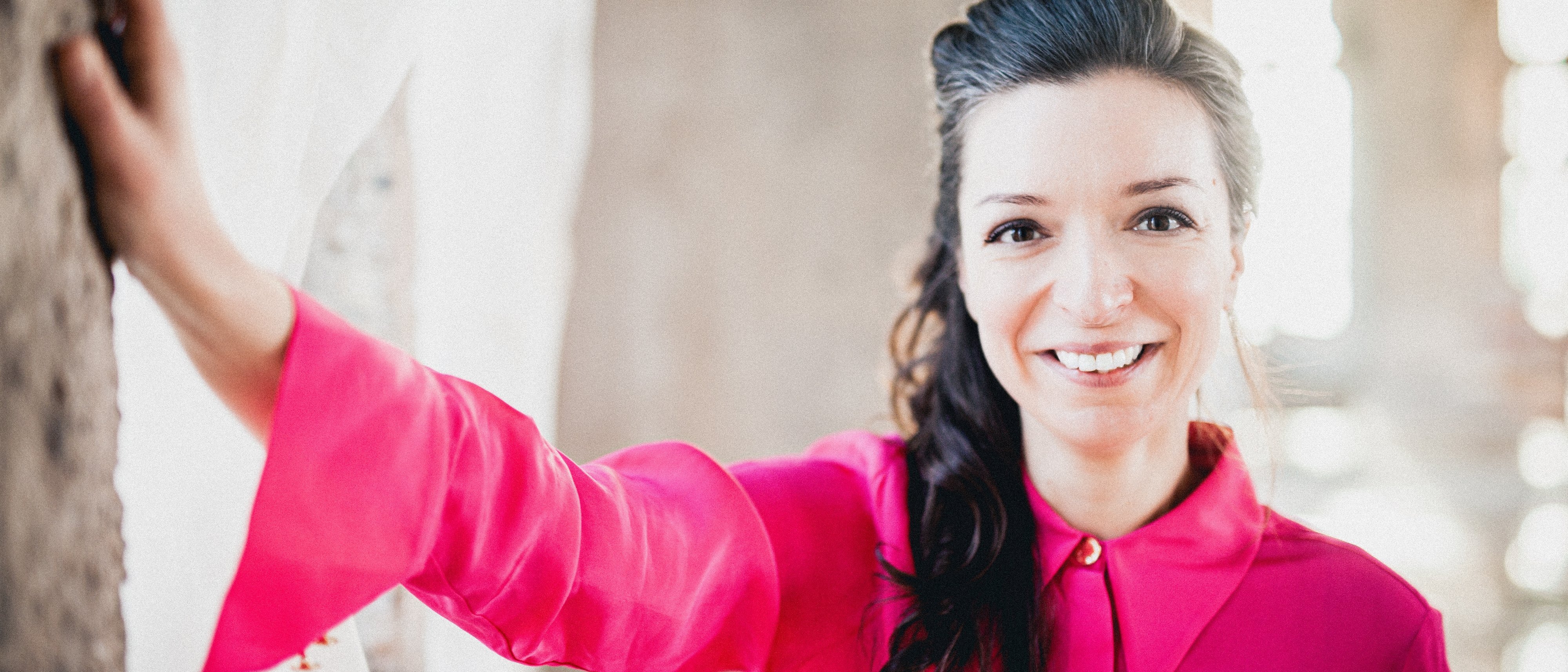 Yulianna Avdeeva, wearing a bright pink blouse, stands indoors and rests her hand on the wall. The background is slightly blurred, which emphasises her cheerful facial expression and the bright colours of her clothes.