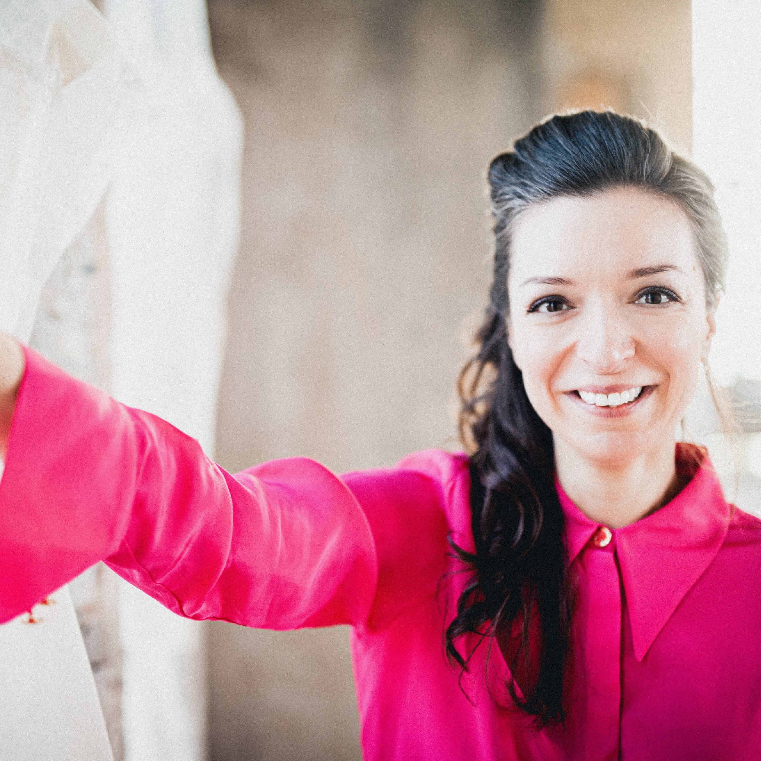Yulianna Avdeeva  in einer leuchtend rosa Bluse steht im Innenbereich und stützt ihre Hand an die Wand. Der Hintergrund ist leicht verschwommen, was ihren fröhlichen Gesichtsausdruck und die leuchtenden Farben der Kleidung hervorhebt.