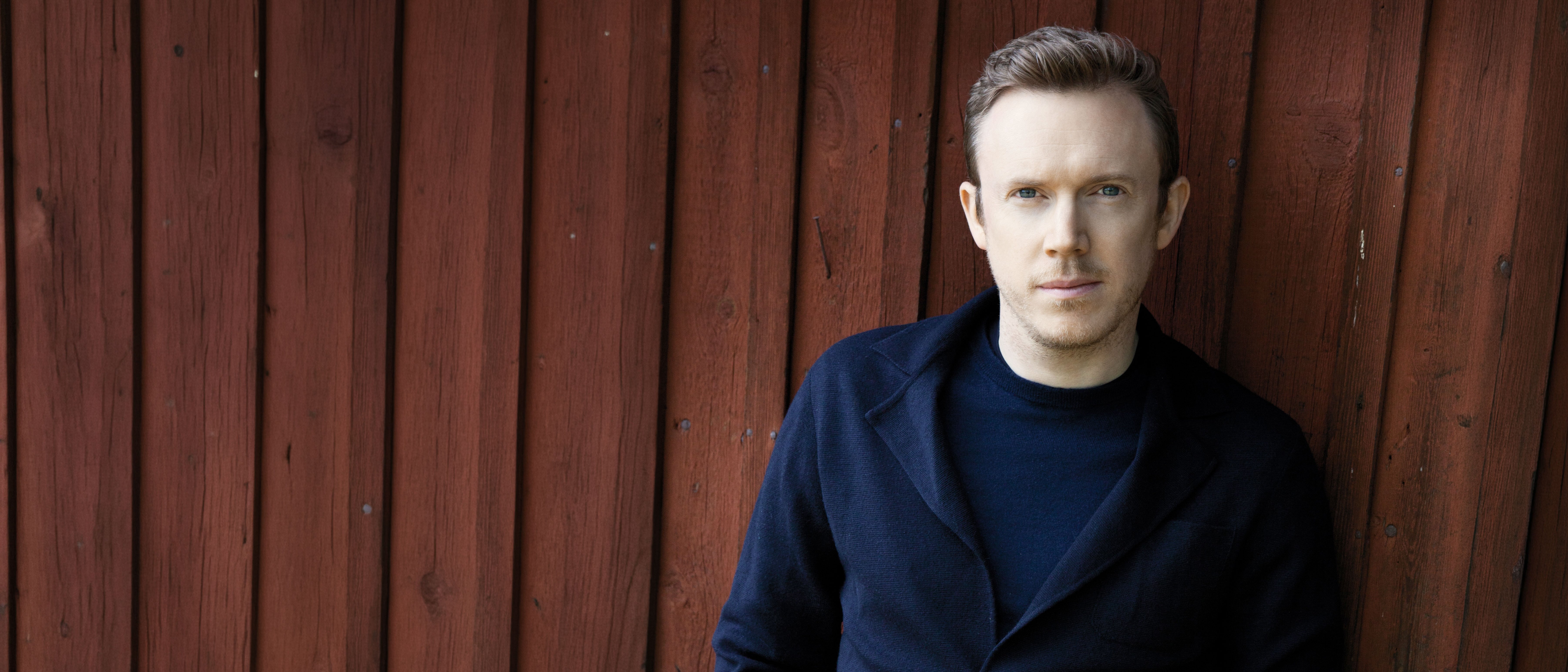 Daniel Harding in einer dunklen Jacke steht mit den Händen in den Taschen an eine rote Holzwand gelehnt und blickt mit neutraler Miene in die Kamera.