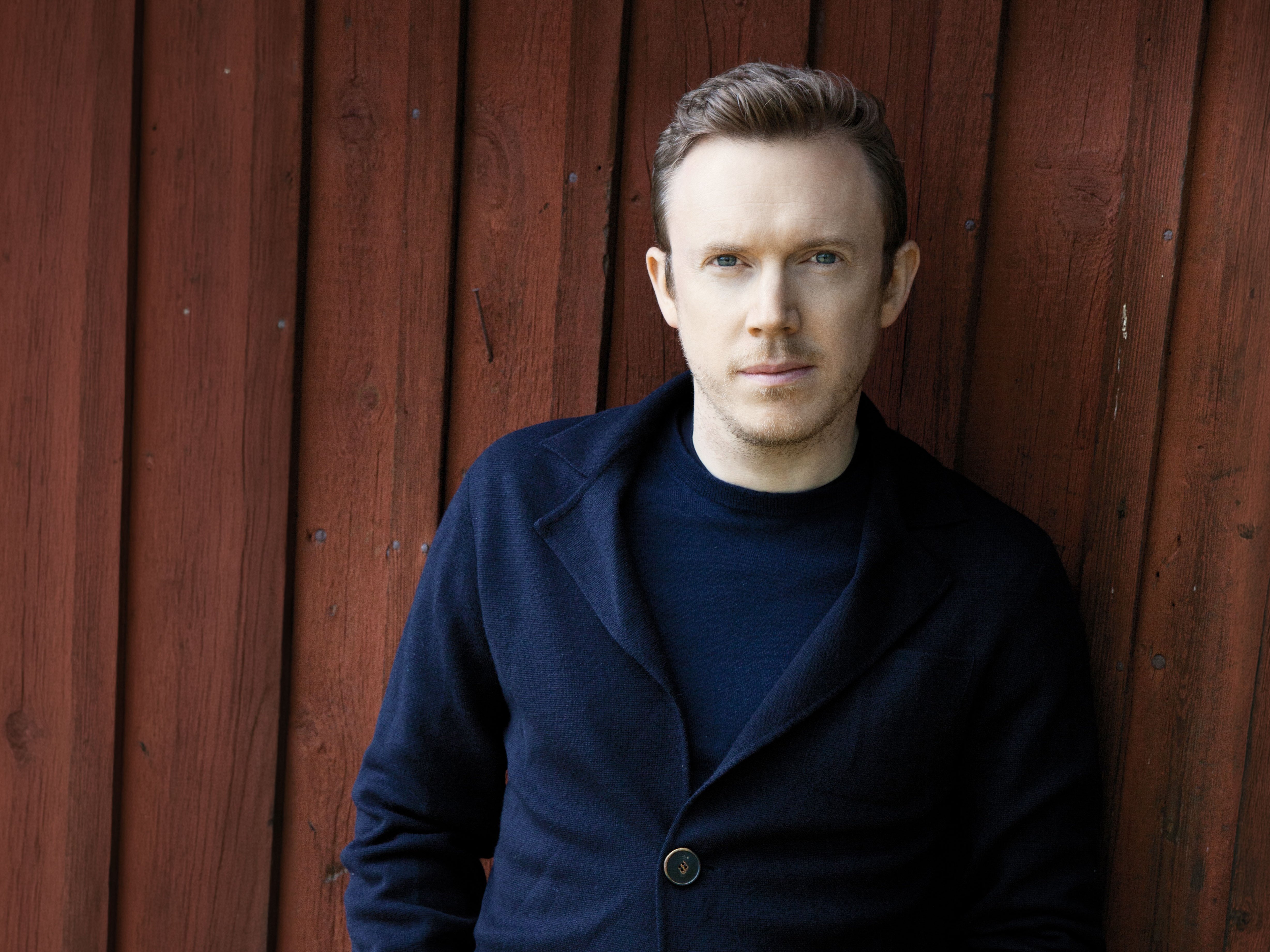 Daniel Harding in einer dunklen Jacke steht mit den Händen in den Taschen an eine rote Holzwand gelehnt und blickt mit neutraler Miene in die Kamera.