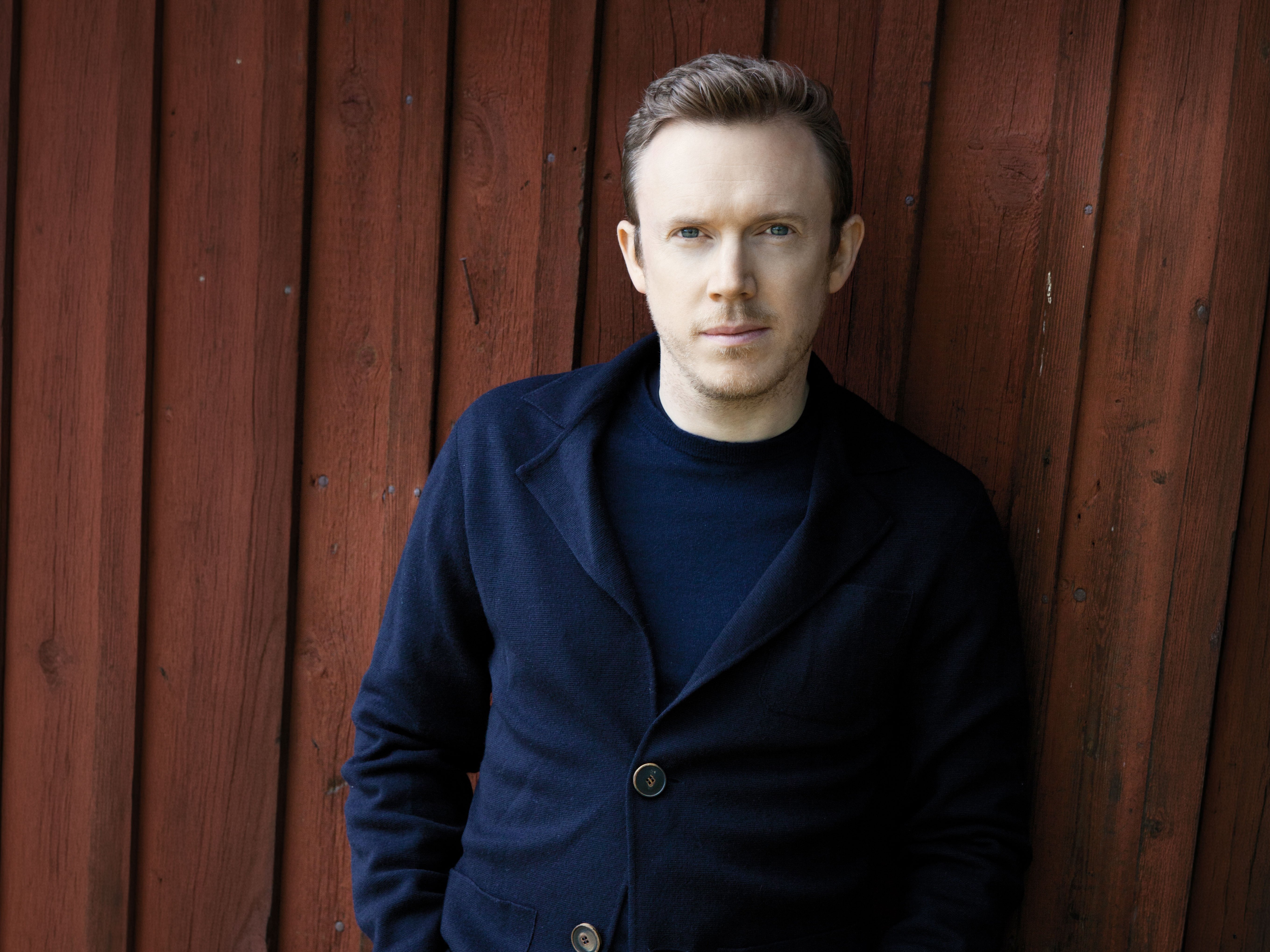 Daniel Harding in a navy jacket and jeans stands against a vertical wooden red wall, looking at the camera with his hands in his pockets. The lighting is soft and the mood is calm and contemplative.