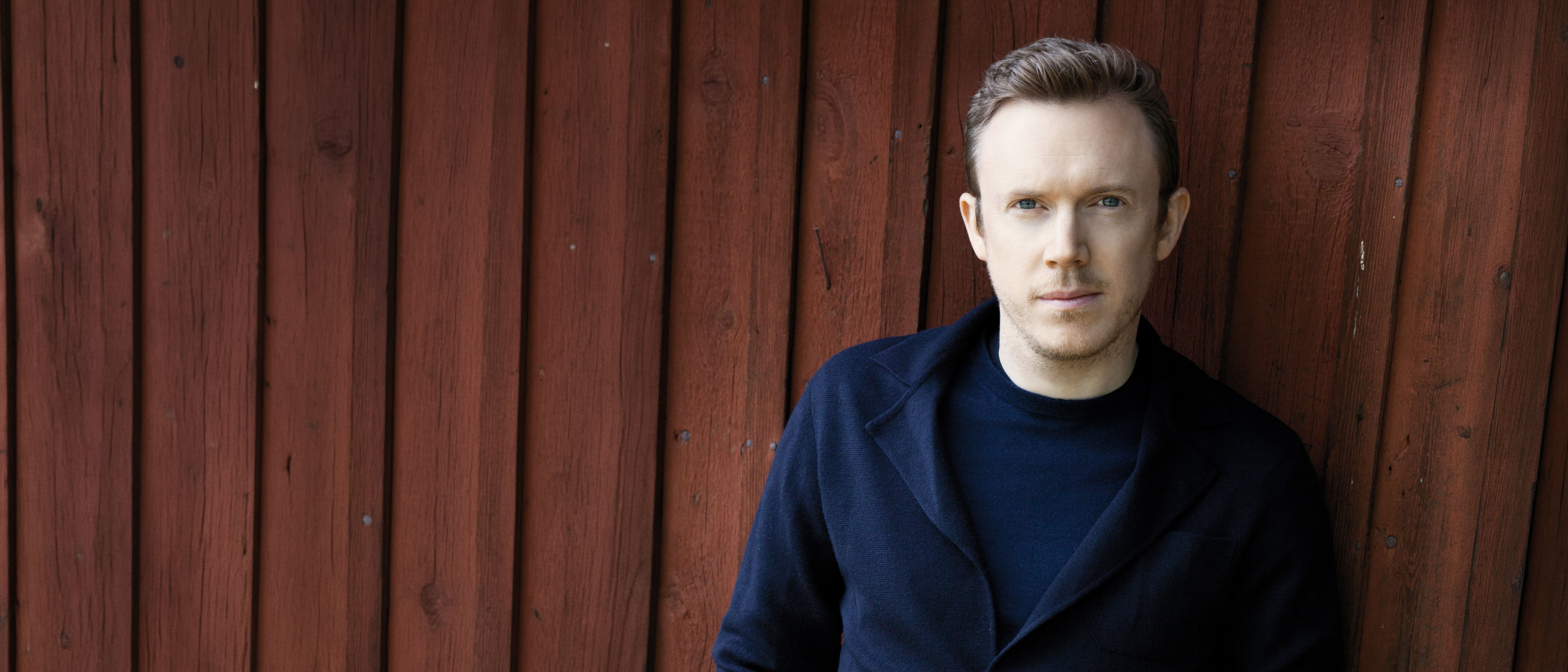 Daniel Harding in einer dunklen Jacke steht mit den Händen in den Taschen an eine rote Holzwand gelehnt und blickt mit neutraler Miene in die Kamera.