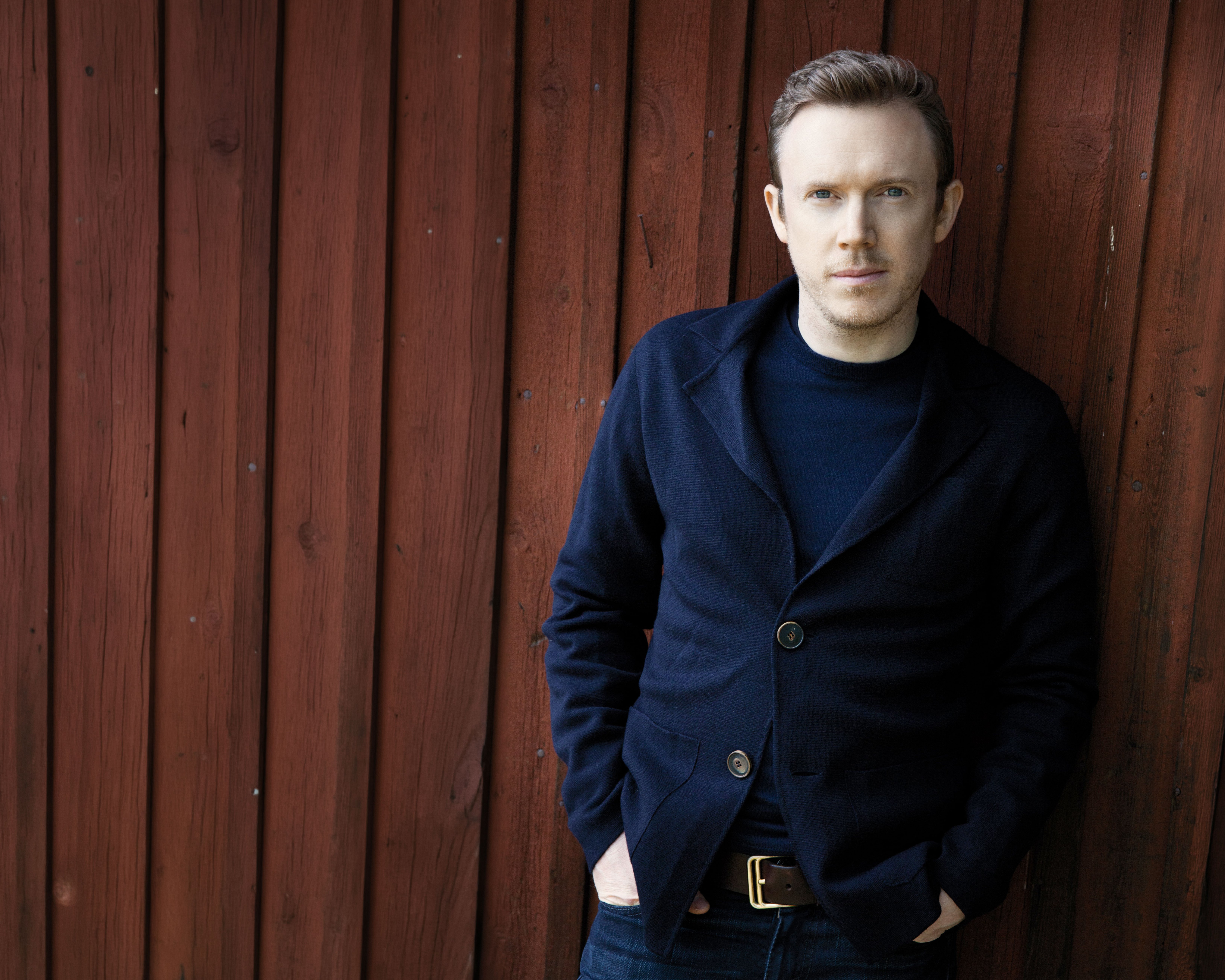 Daniel Harding in einer dunklen Jacke steht mit den Händen in den Taschen an eine rote Holzwand gelehnt und blickt mit neutraler Miene in die Kamera.