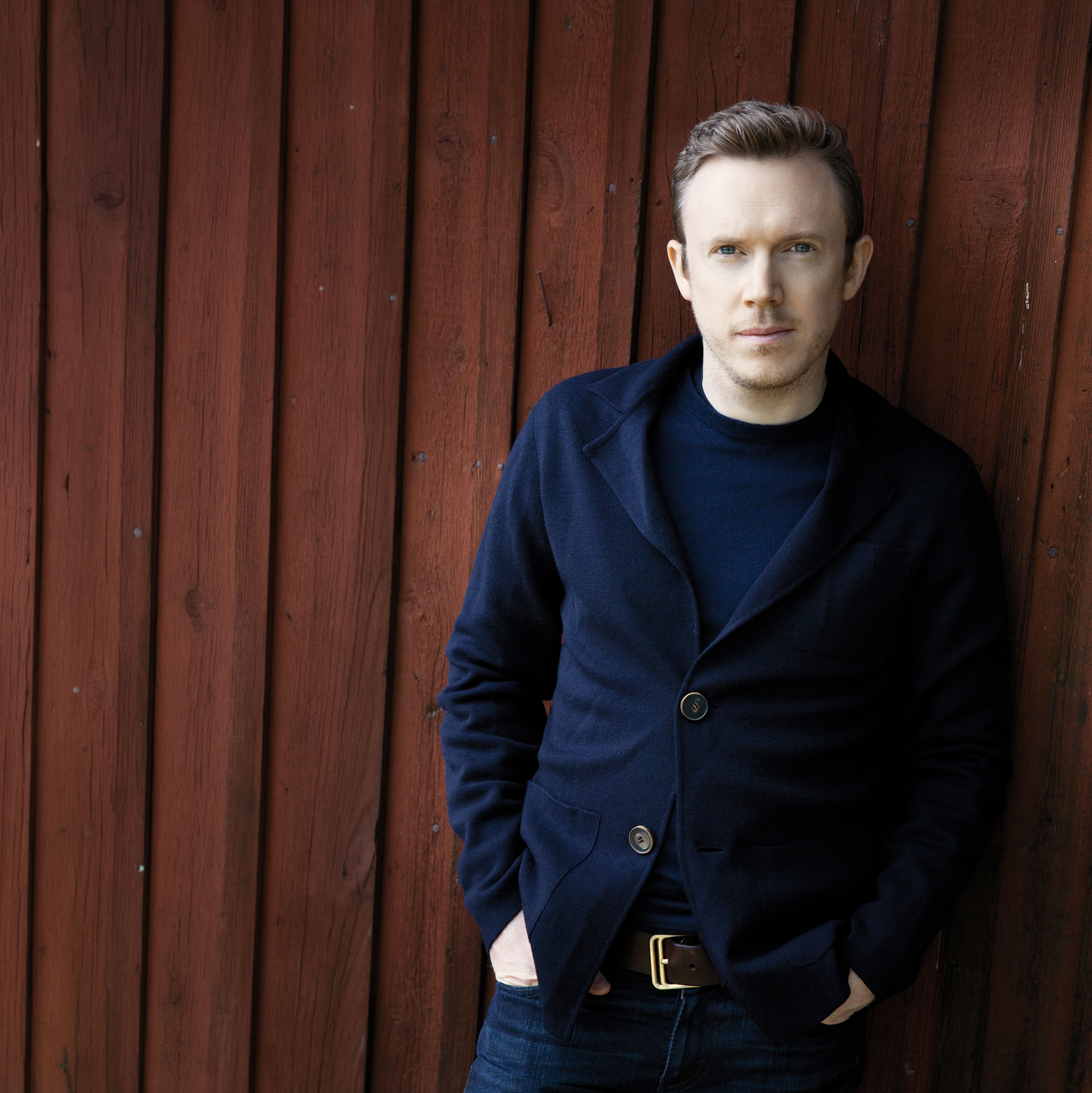 Daniel Harding in einer dunklen Jacke steht mit den Händen in den Taschen an eine rote Holzwand gelehnt und blickt mit neutraler Miene in die Kamera.