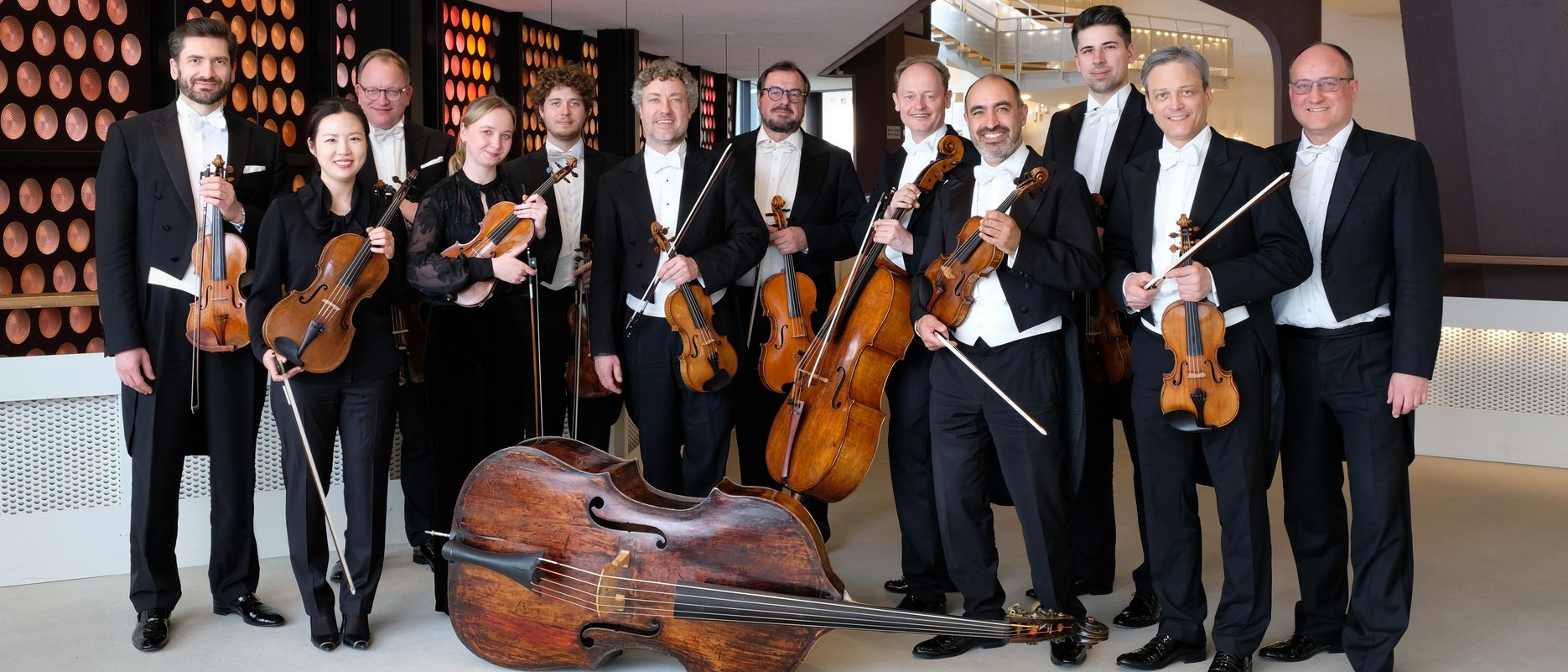 Eine Gruppe von Musikern in formeller schwarzer Kleidung posiert in einem Innenbereich mit Streichinstrumenten, darunter Geigen, Bratschen, Celli und einem Kontrabass, vor einer modernen architektonischen Kulisse.