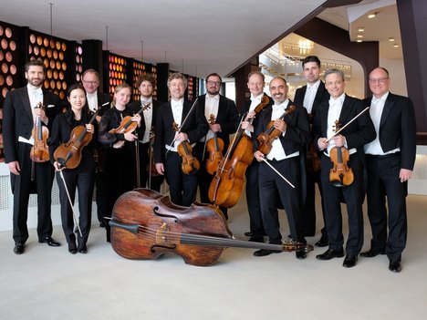 Eine Gruppe von Musikern in formeller schwarzer Kleidung posiert in einem Innenbereich mit Streichinstrumenten, darunter Geigen, Bratschen, Celli und einem Kontrabass, vor einer modernen architektonischen Kulisse.