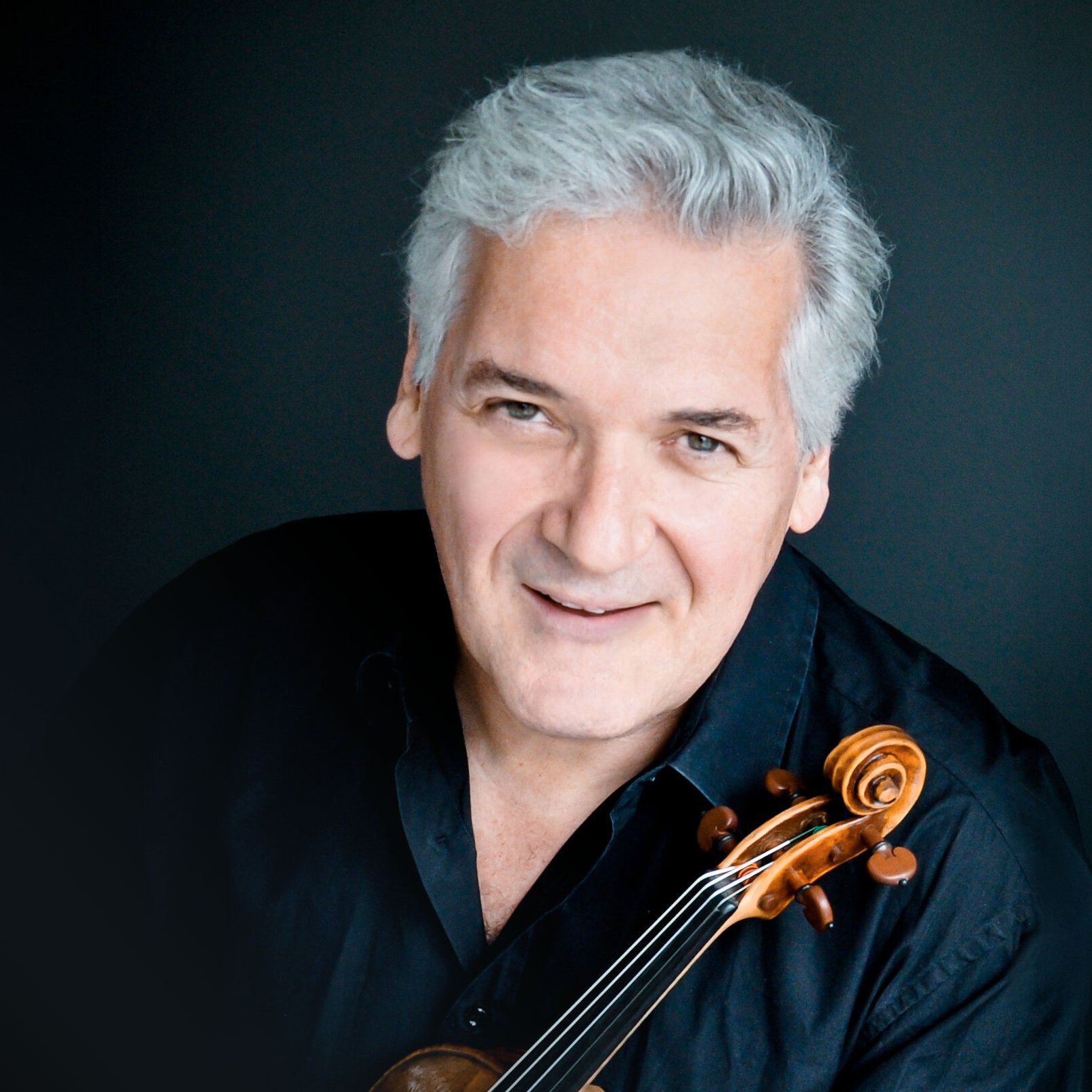 Ein lächelnder Pinchas Zukerman mit grauem Haar und schwarzem Hemd posiert mit einer Geige vor einem dunklen Hintergrund.