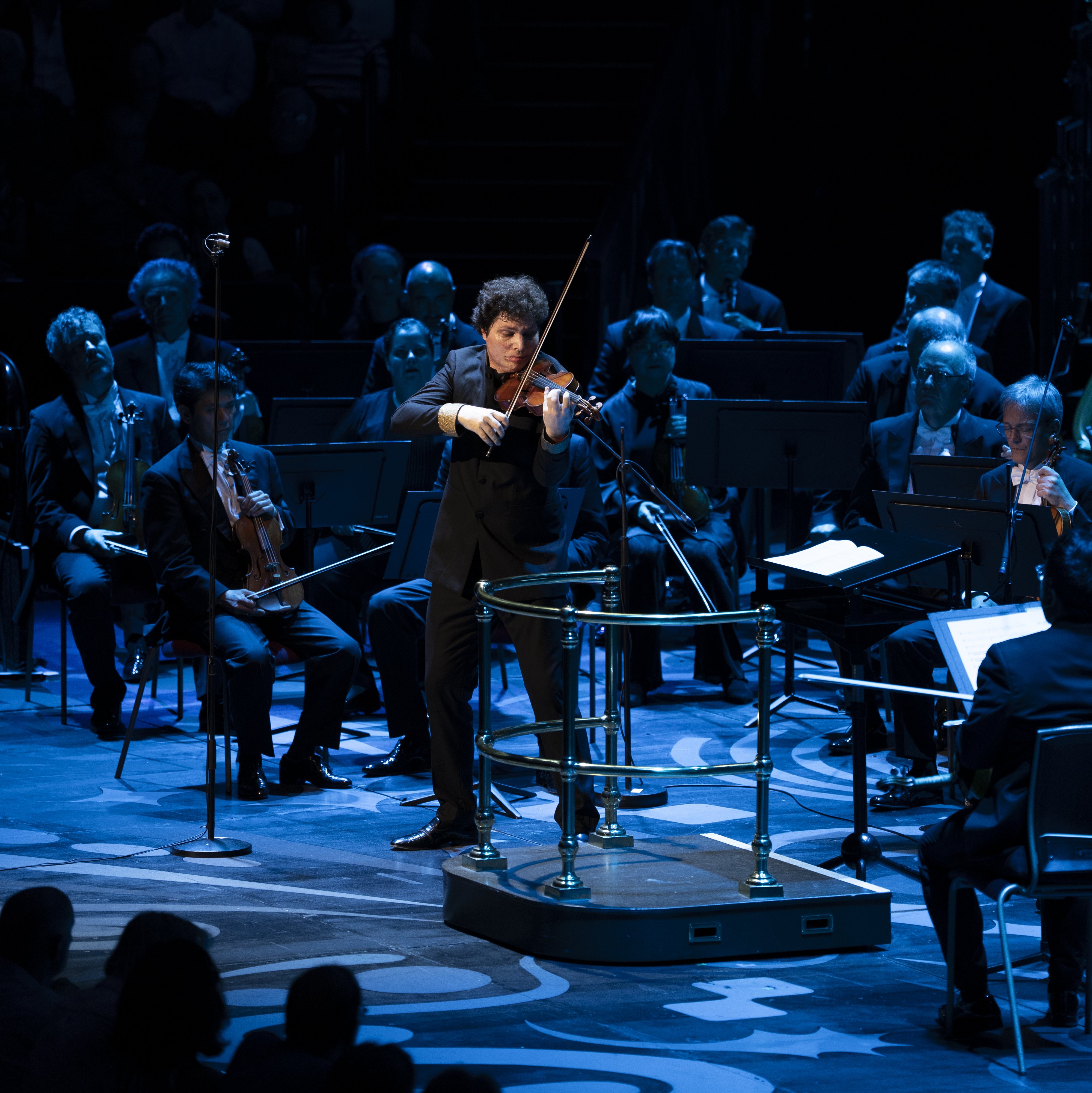 A solo violinist performs on stage with an orchestra seated behind, under dramatic blue lighting, while an audience watches in the dimly lit foreground.