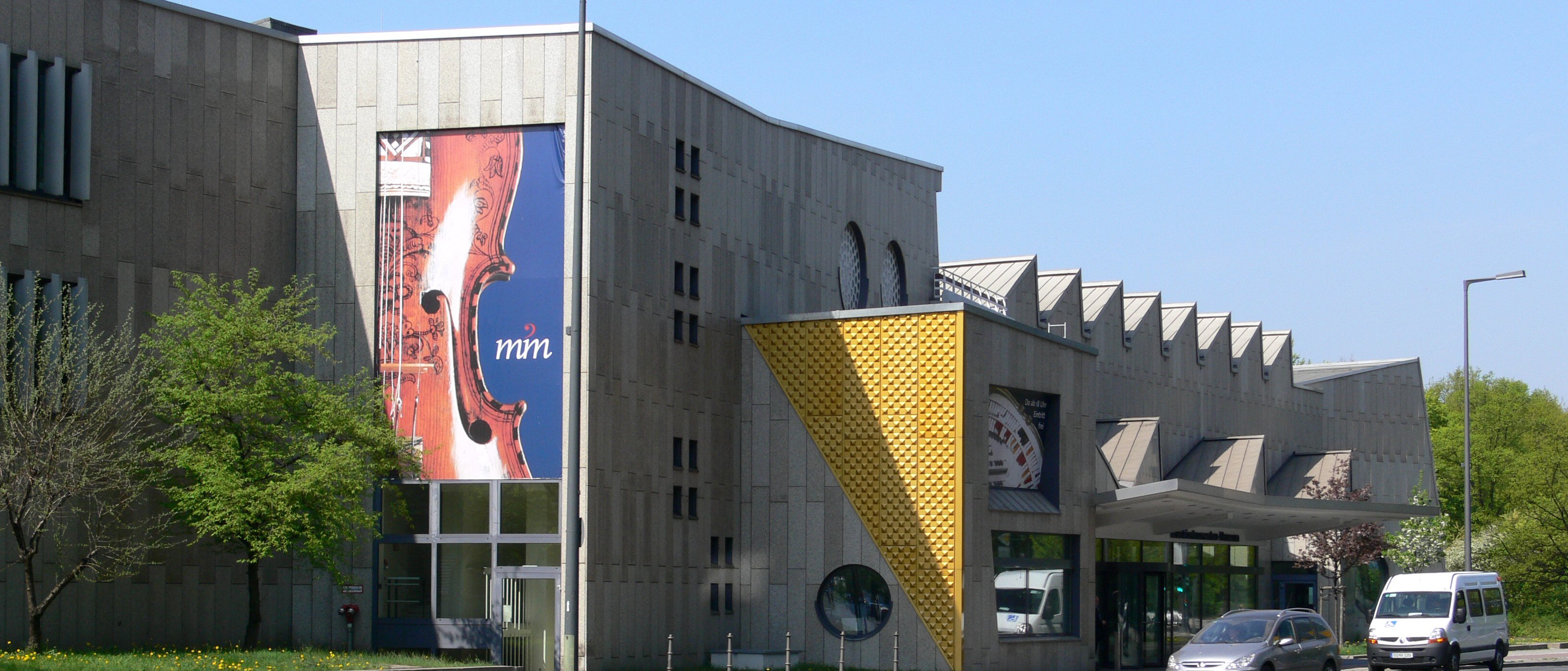 Ein modernes Betongebäude mit geometrischen Formen und großen Fenstern, an dessen Fassade ein Banner mit dem Thema Geige prangt. Auf der Straße davor parken und fahren Autos. In der Nähe gibt es Bäume und Grünflächen.