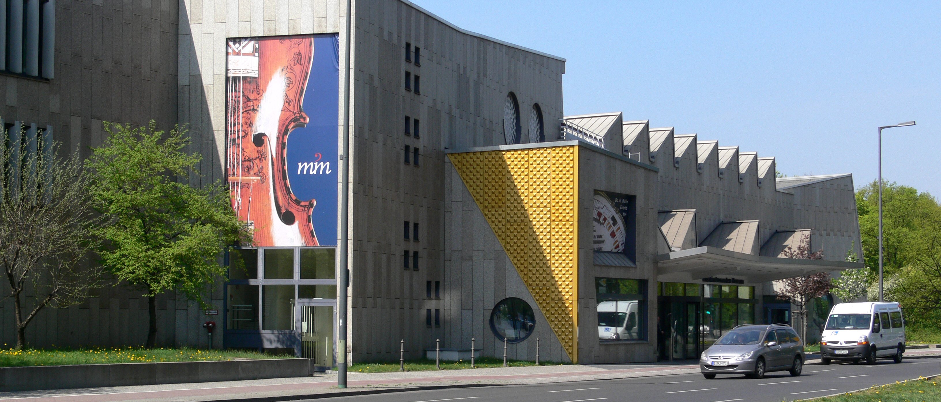 Ein modernes Betongebäude mit geometrischen Formen und großen Fenstern, an dessen Fassade ein Banner mit dem Thema Geige prangt. Auf der Straße davor parken und fahren Autos. In der Nähe gibt es Bäume und Grünflächen.
