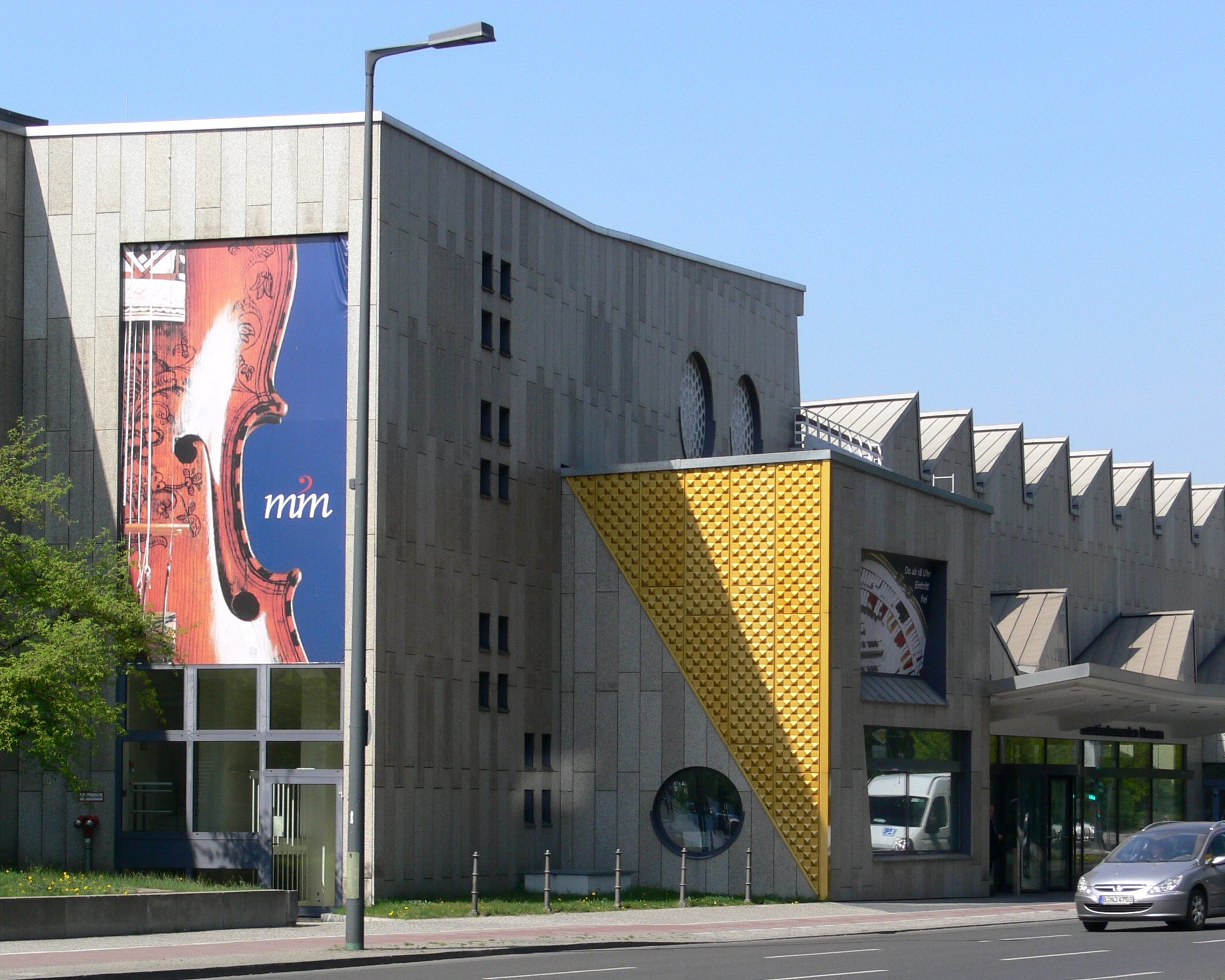 Ein modernes Betongebäude mit geometrischen Formen und großen Fenstern, an dessen Fassade ein Banner mit dem Thema Geige prangt. Auf der Straße davor parken und fahren Autos. In der Nähe gibt es Bäume und Grünflächen.