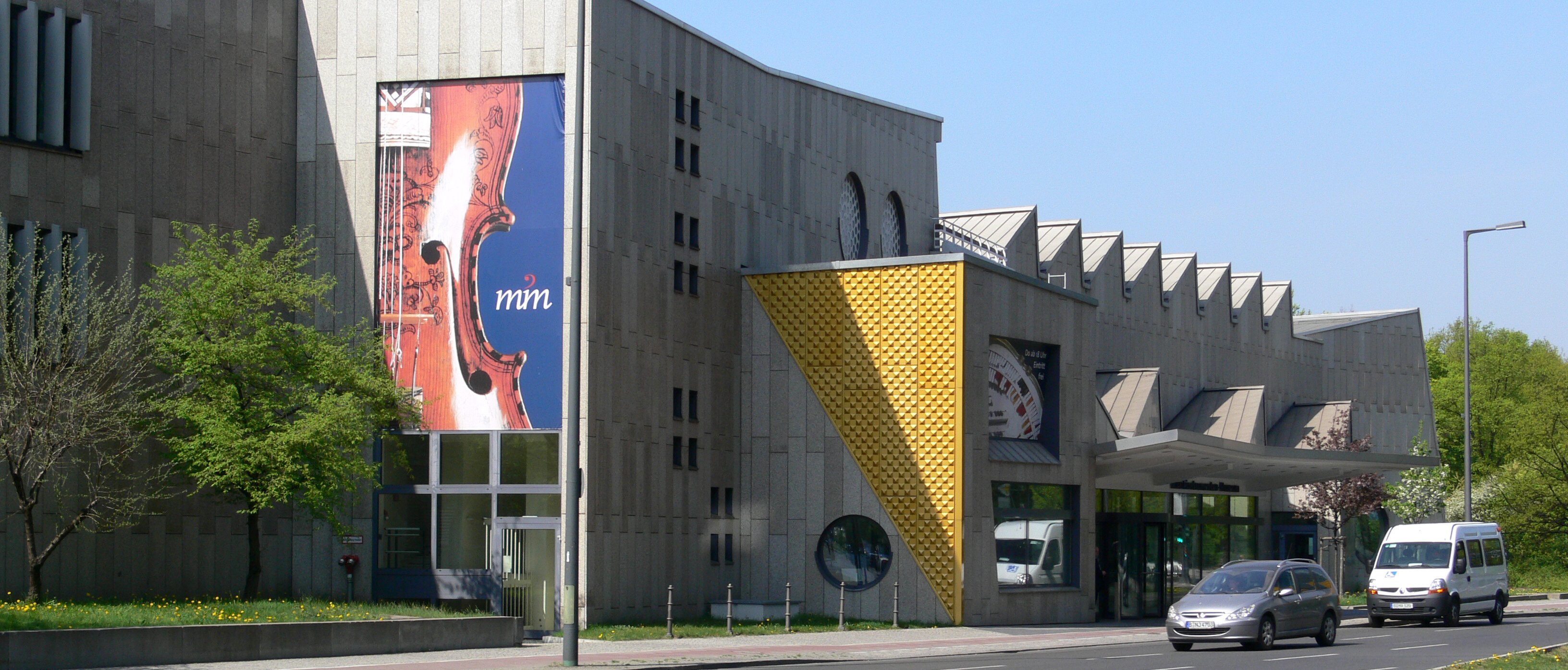 Ein modernes Betongebäude mit geometrischen Formen und großen Fenstern, an dessen Fassade ein Banner mit dem Thema Geige prangt. Auf der Straße davor parken und fahren Autos. In der Nähe gibt es Bäume und Grünflächen.
