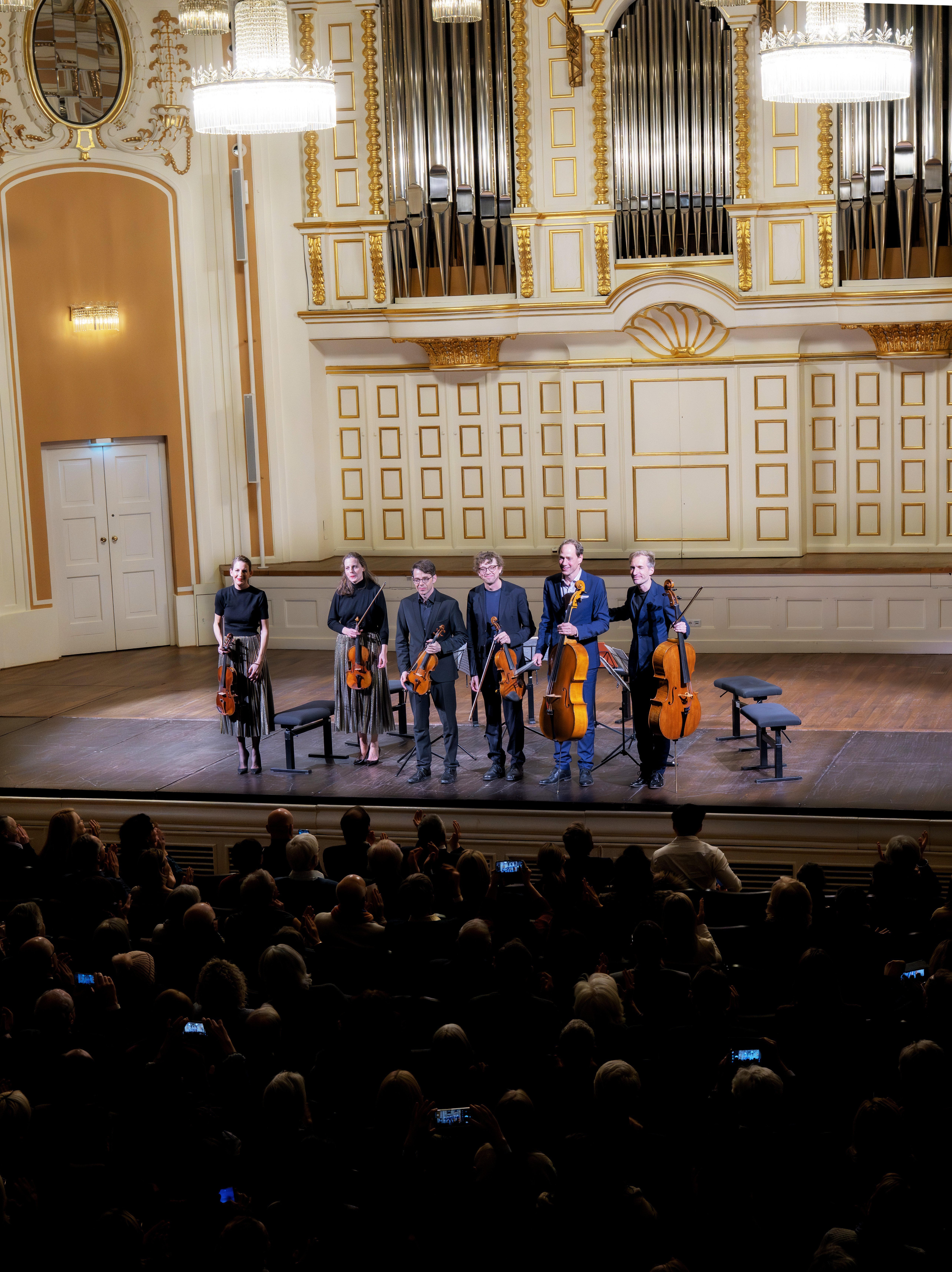 Sechs Musiker mit Streichinstrumenten stehen auf der Bühne und verbeugen sich in einem prunkvollen Konzertsaal vor einem im Vordergrund sitzenden Publikum. Hinter ihnen sind große Orgelpfeifen und Kronleuchter zu sehen.