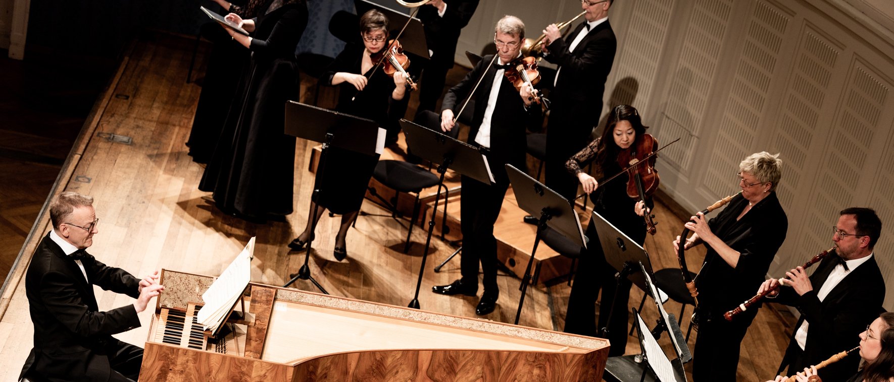 A chamber orchestra performs on stage, featuring string and woodwind musicians, with a harpsichordist seated at the keyboard in the foreground. Music stands and sheet music are visible.