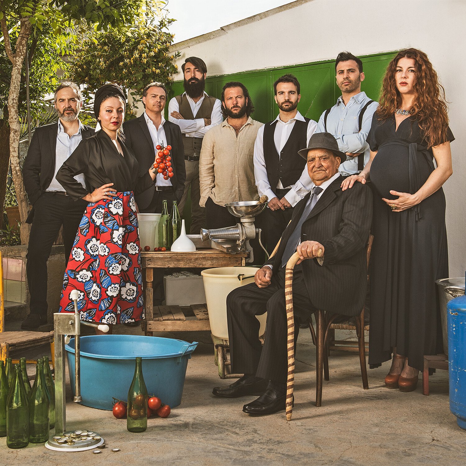 Neun Erwachsene posieren gemeinsam im Freien mit Kisten, Flaschen, Schüsseln und einem blauen Benzintank. Eine Frau ist schwanger, ein Mann sitzt mit einem Stock und die anderen stehen in verschiedenen Posen und sind halb-formal gekleidet.