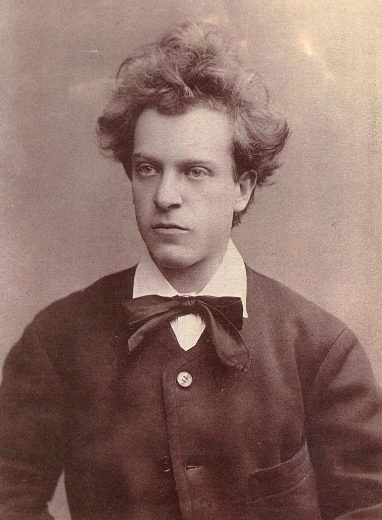 Sepia-toned portrait of a young man with wavy, disheveled hair, wearing a dark jacket, white shirt, and a large bow tie, looking slightly to the side with a serious expression.