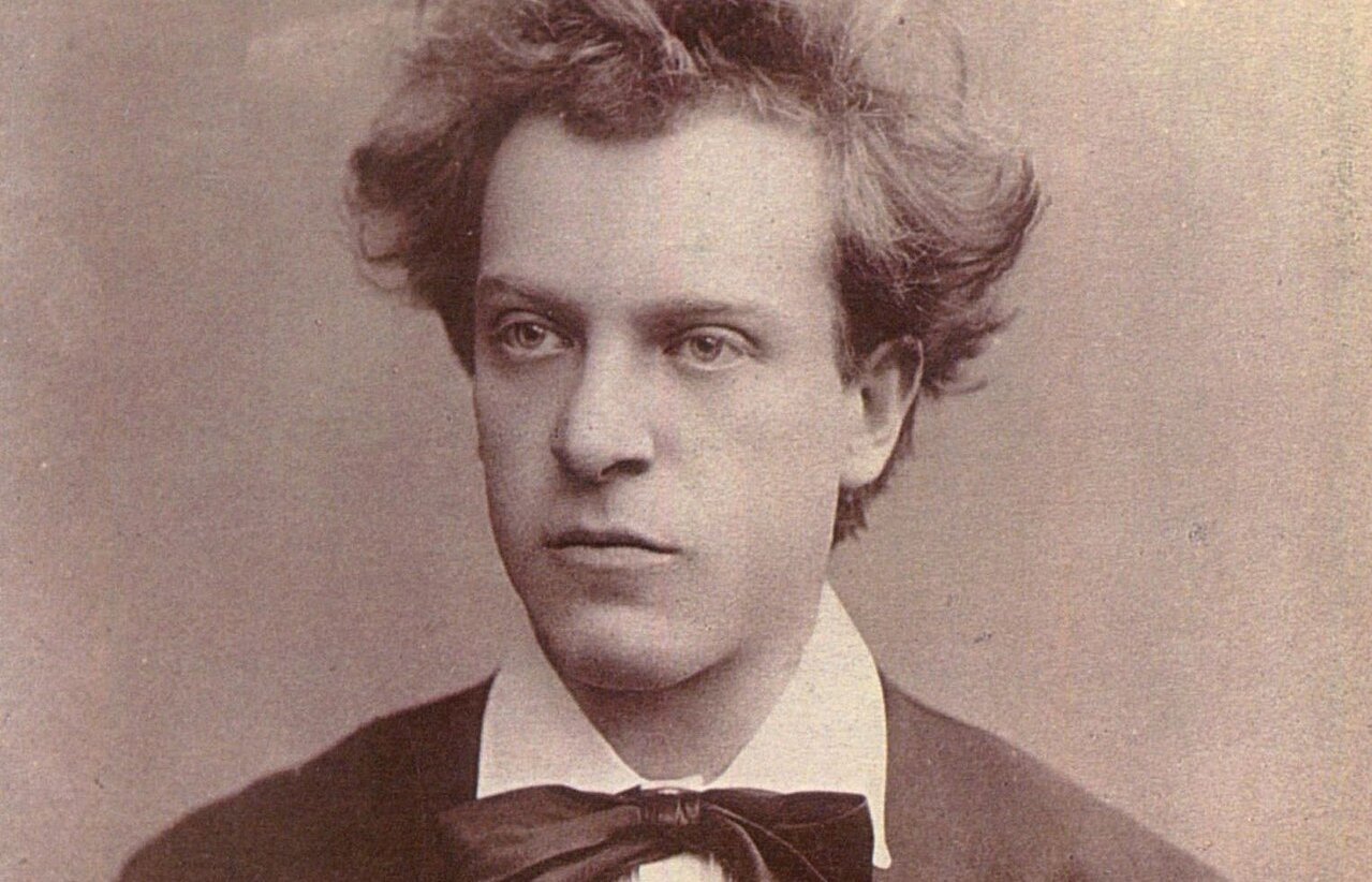 Sepiafarbenes Porträt eines jungen Mannes mit gewelltem, zerzaustem Haar, der eine dunkle Jacke, ein weißes Hemd und eine große Fliege trägt und mit einem neutralen Gesichtsausdruck leicht zur Seite blickt.