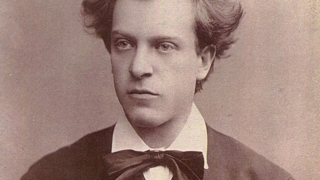 Sepia-toned portrait of a young man with wavy, disheveled hair, wearing a dark jacket, white shirt, and a large bow tie, looking slightly to the side with a serious expression.