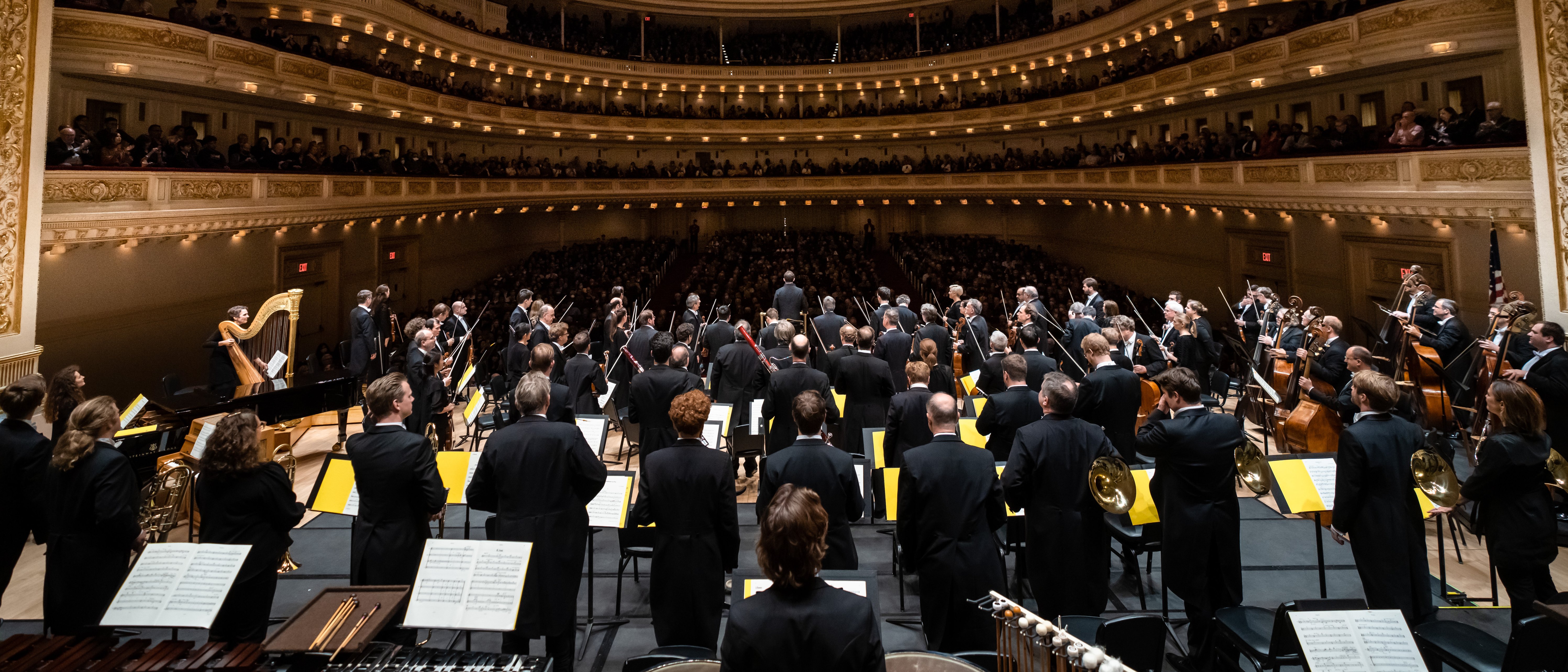 A full orchestra stands on stage in a grand concert hall, facing an audience filling multiple balconies, with elegant chandeliers overhead and ornate gold detailing throughout the space.