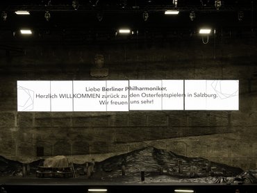 A stage with technical equipment and crew in the foreground. A large screen above the stage displays a German welcome message to the Berliner Philharmoniker at the Salzburg Easter Festival.