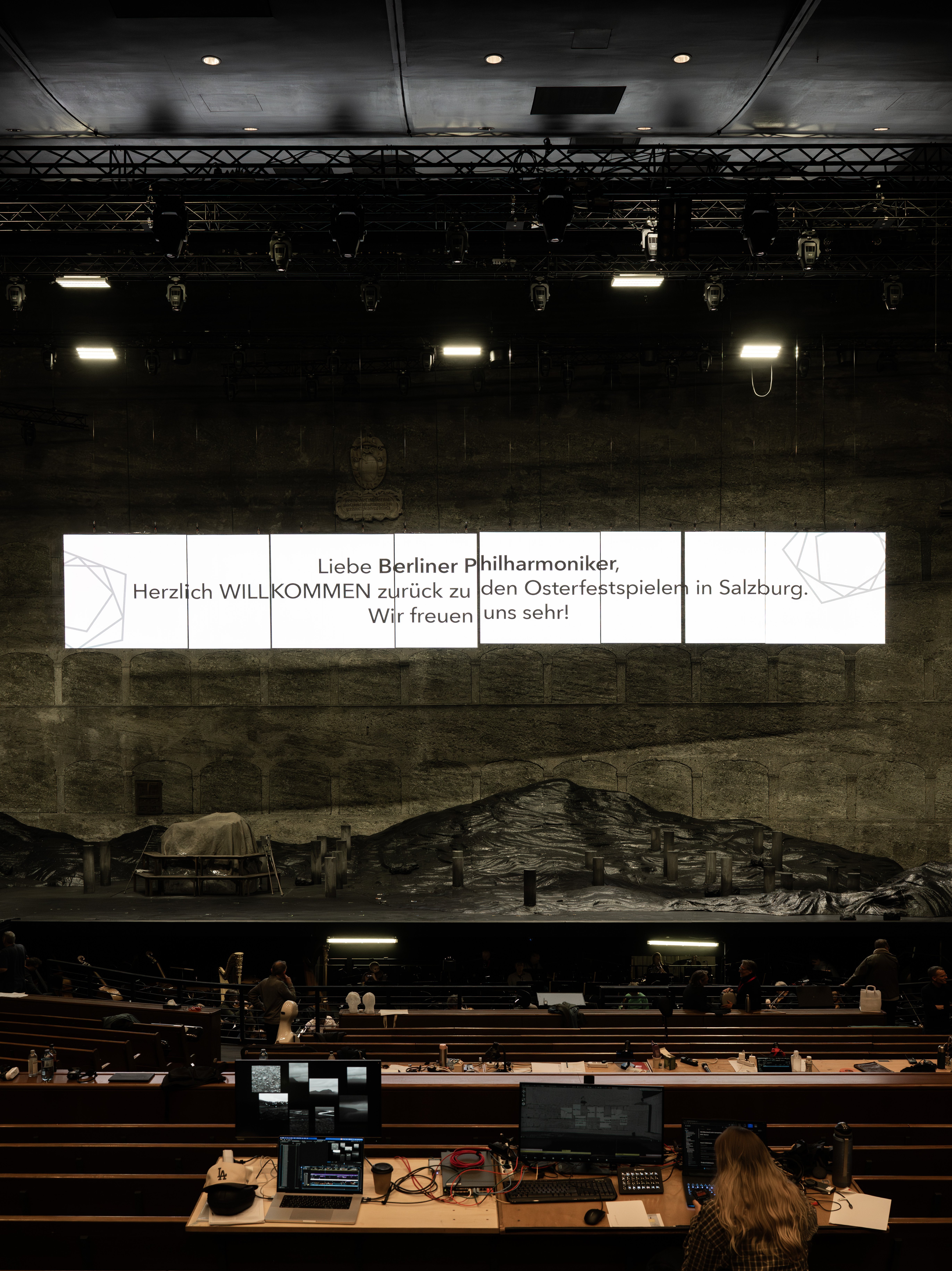 Eine Konzertsaalbühne mit einem großen Willkommensgruß in deutscher Sprache darüber, im schwach beleuchteten Vordergrund sind Techniker und Geräte zu sehen.
