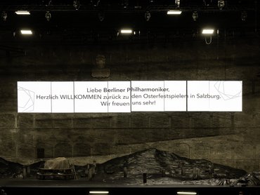 Eine Konzertsaalbühne mit einem großen Willkommensgruß in deutscher Sprache darüber, im schwach beleuchteten Vordergrund sind Techniker und Geräte zu sehen.