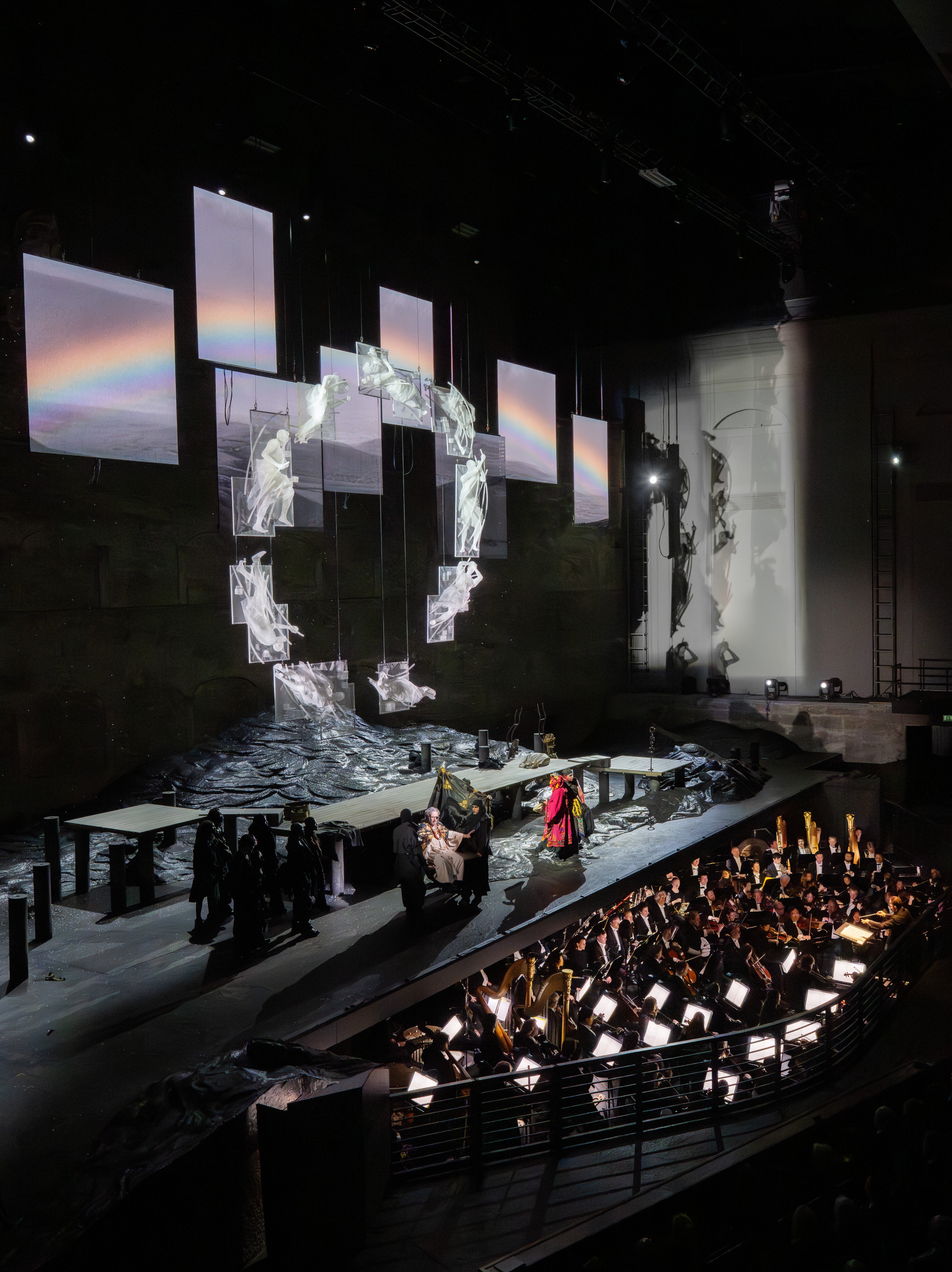 Ein Orchester spielt unter einem dramatischen Bühnenbild mit digitalen Bildschirmen, felsigem Gelände und Darstellern in Kostümen bei theatralischer Beleuchtung in einem großen Theater.