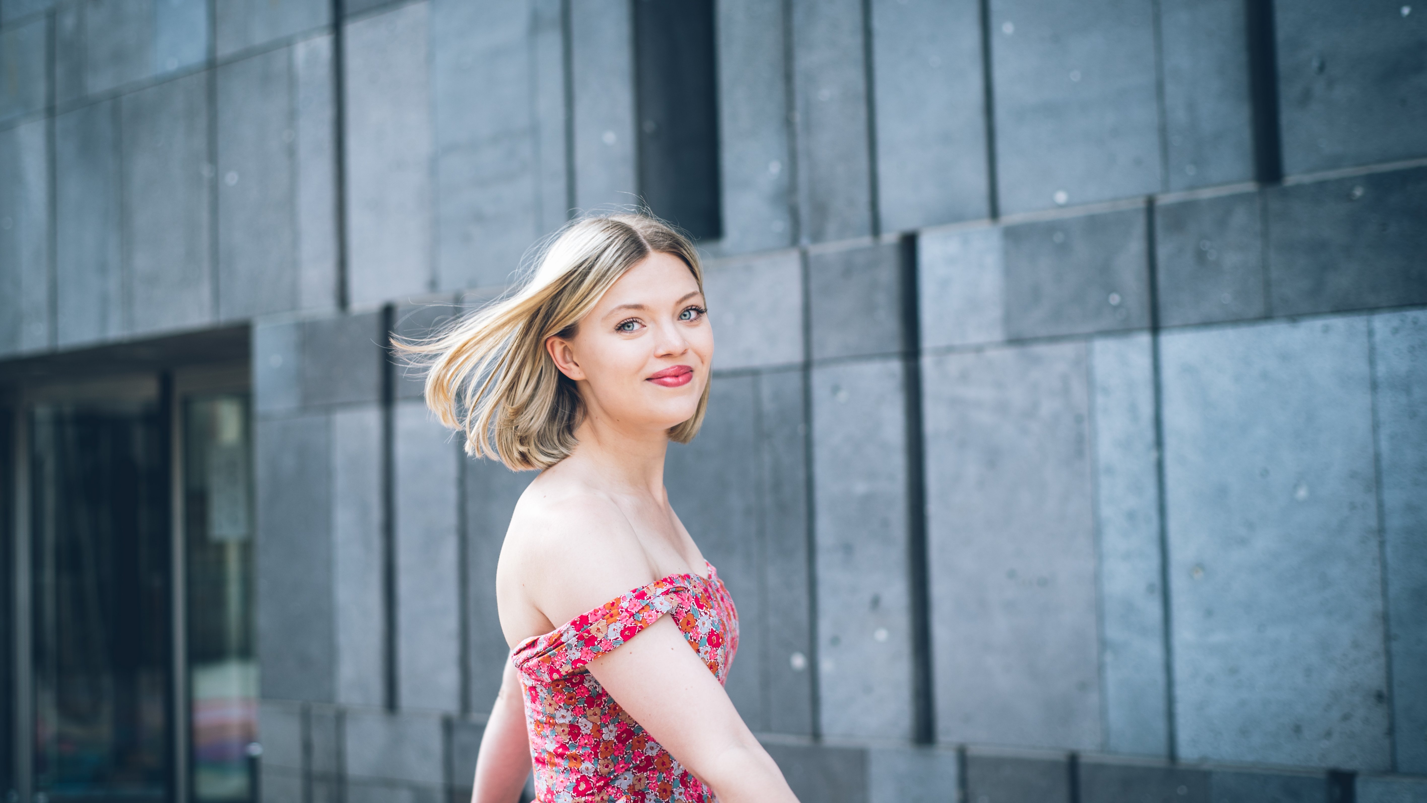 Eine lächelnde Frau in einem geblümten, schulterfreien Kleid steht im Freien vor einer modernen grauen Steinmauer; ihr blondes Haar weht sanft im Wind, während sie sich der Kamera zuwendet.
