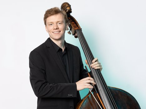A young man in a black suit smiles while playing an upright bass against a plain white background. He holds the instrument with both hands and looks at the camera.
