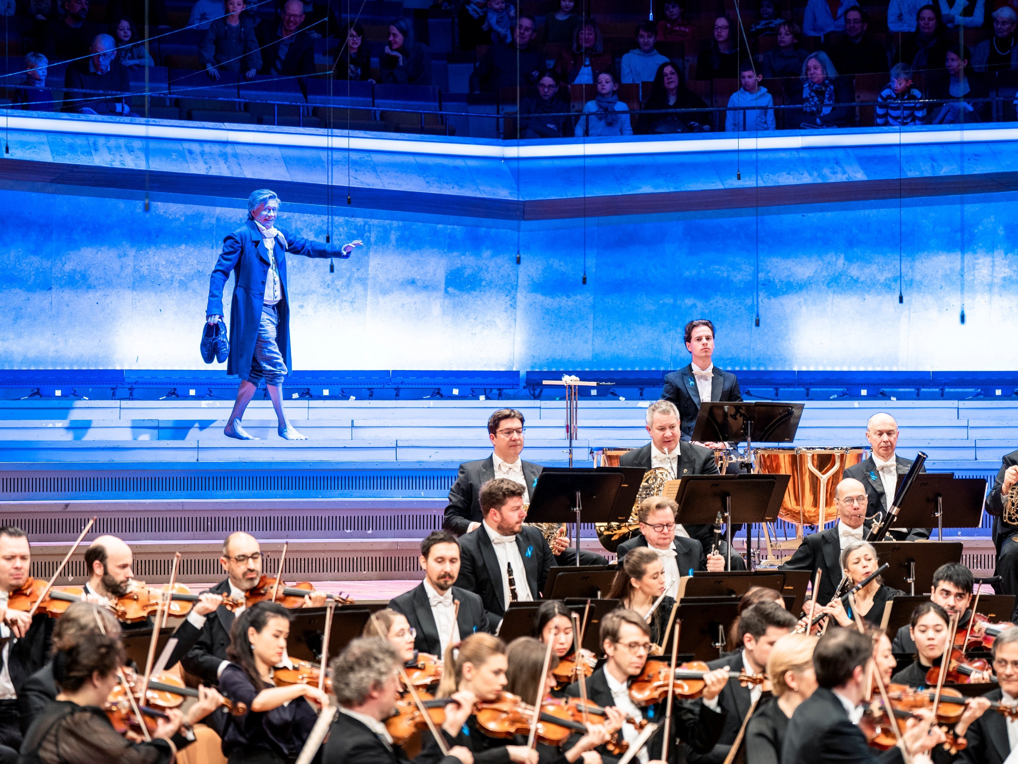 Ein Orchester spielt auf der Bühne, während ein Mann in einem blauen langen Mantel auf beleuchteten Stufen hinter ihnen steht und mit dem Arm gestikuliert. Im Hintergrund sind die Zuschauer zu sehen.