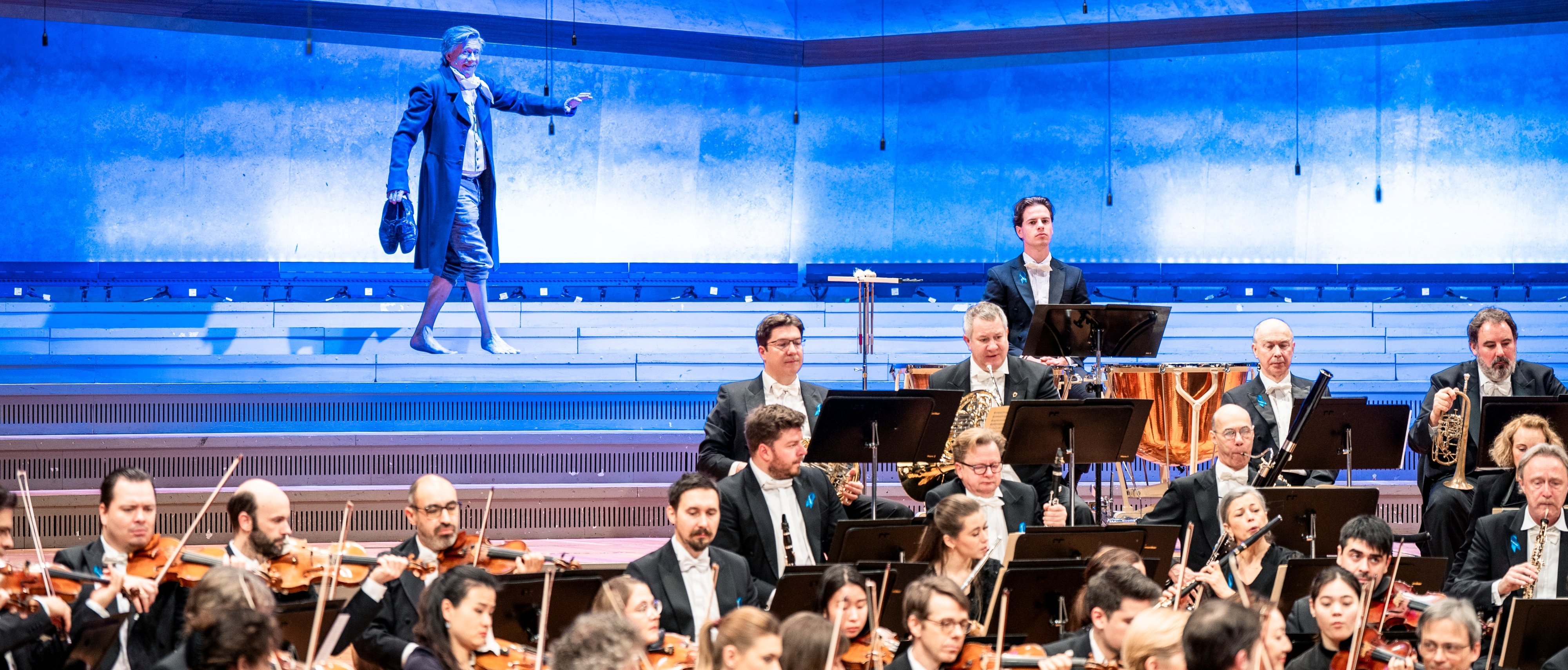 Ein Orchester spielt auf der Bühne, während ein Mann in einem blauen langen Mantel auf beleuchteten Stufen hinter ihnen steht und mit dem Arm gestikuliert. Im Hintergrund sind die Zuschauer zu sehen.