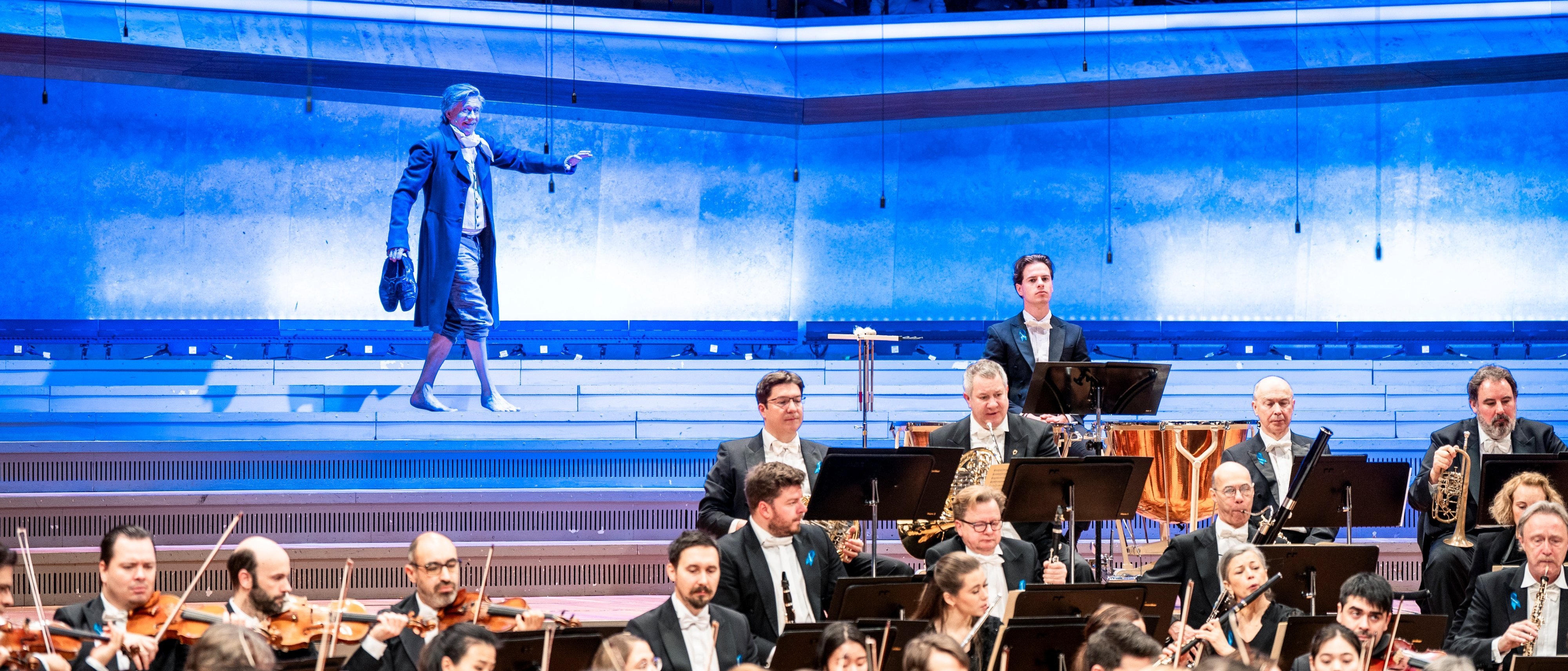 Ein Orchester spielt auf der Bühne, während ein Mann in einem blauen langen Mantel auf beleuchteten Stufen hinter ihnen steht und mit dem Arm gestikuliert. Im Hintergrund sind die Zuschauer zu sehen.