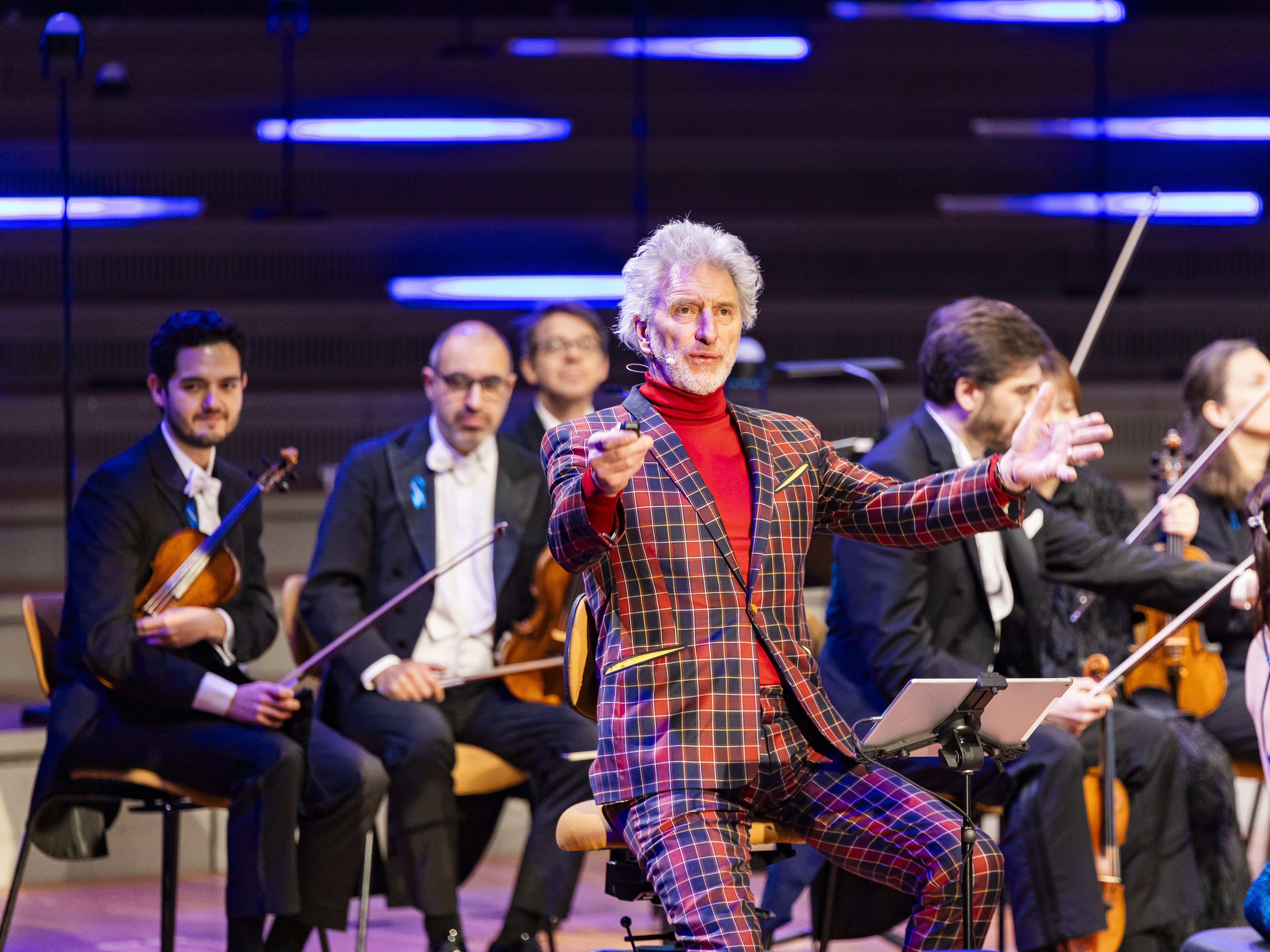 A conductor in a red plaid suit leads an orchestra on stage as musicians with string instruments watch, seated in front of illuminated blue lights in the background.