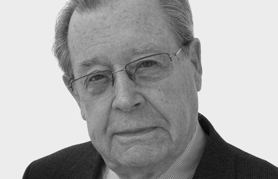 Ein älterer Mann mit Brille, grauem Haar und ernster Miene, der einen dunklen Anzug, ein gestreiftes Hemd und eine gemusterte Krawatte trägt, posiert vor einem schlichten hellen Hintergrund.