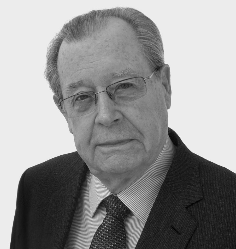 Ein älterer Mann mit Brille, grauem Haar und ernster Miene, der einen dunklen Anzug, ein gestreiftes Hemd und eine gemusterte Krawatte trägt, posiert vor einem schlichten hellen Hintergrund.